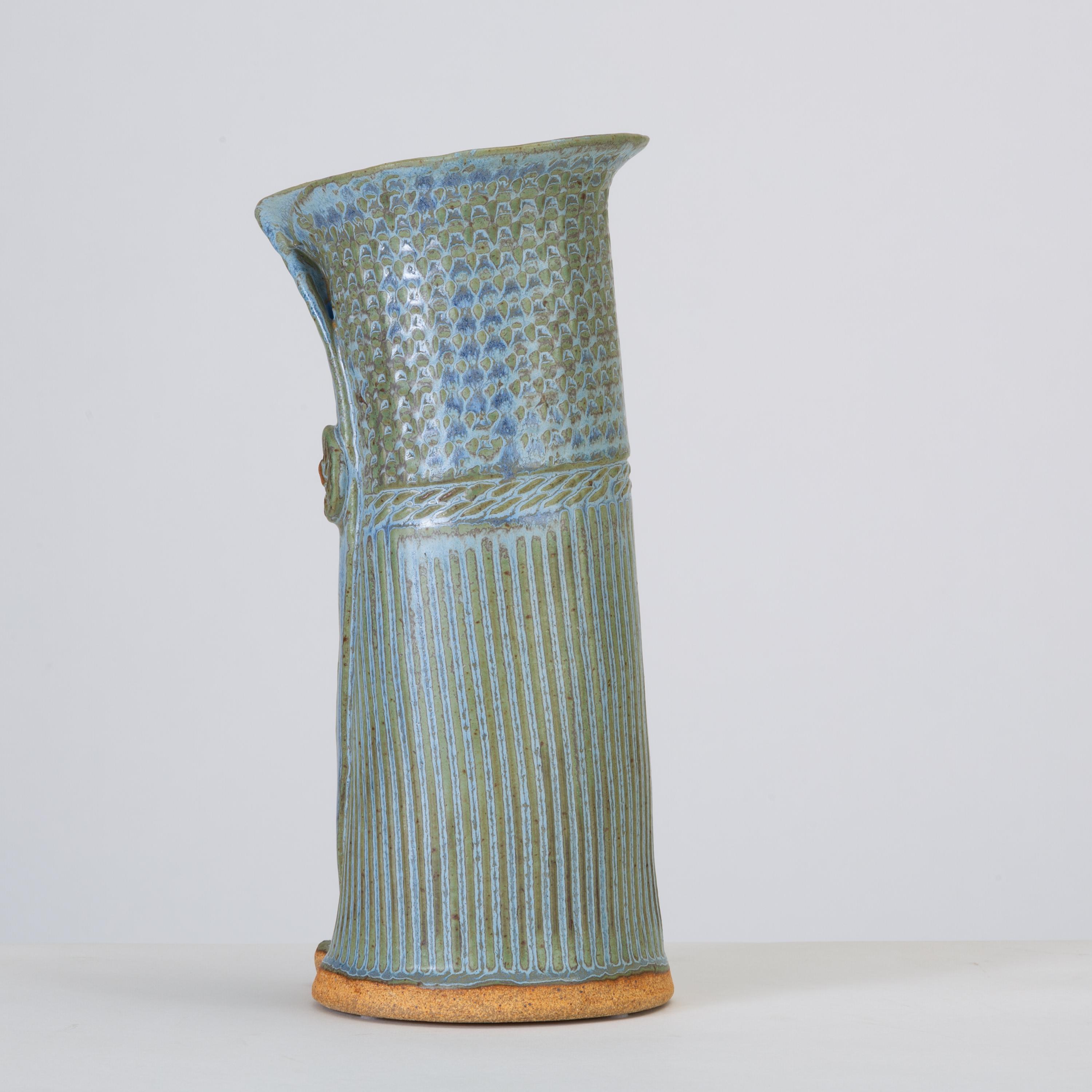 A unique asymmetric vase with a blue-green varicolor glaze and sgraffito detailing. The piece is painted and incised to mimic the fibers of woven textiles and the edges of the clay have been folded over each other to mimic layers of cloth secured by