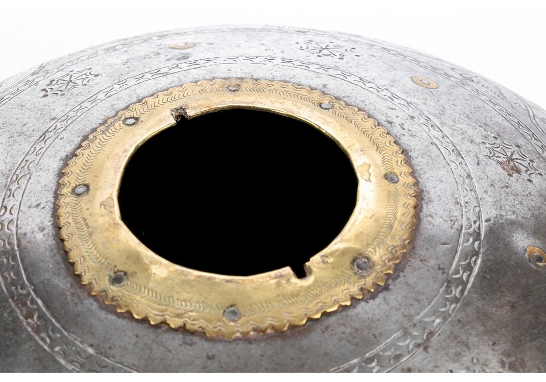A very decorative piece in a flattened form in gray metal with a small top opening rimmed in brass. The lid with brass top with a ring. The jar with chased leafy band on the side and brass roundels on the band above. 

Dimensions: Diameter 13 1/2