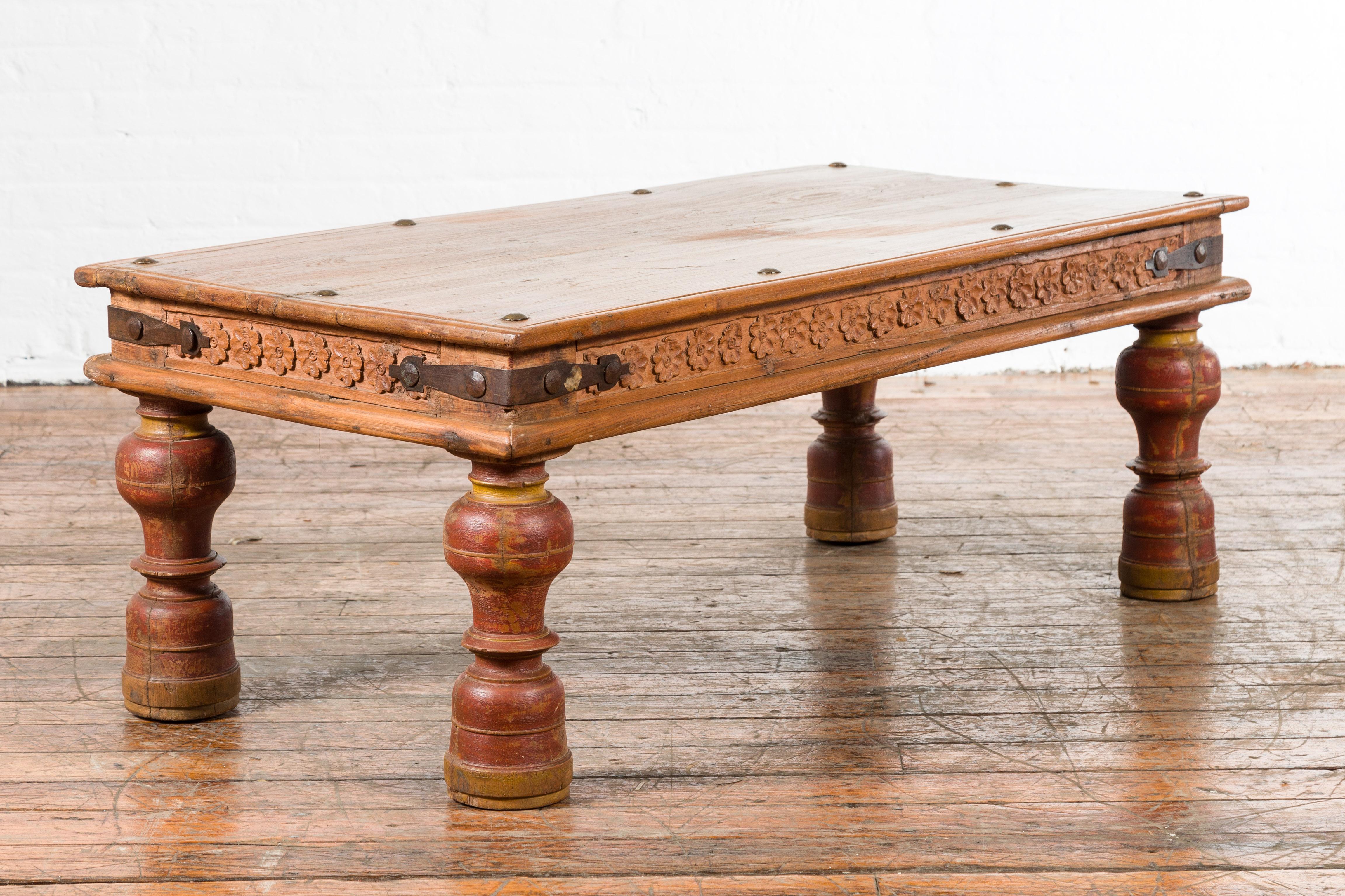 An Indian antique coffee table from the 19th century, with carved floral frieze, baluster legs and iron braces. Created in India during the 19th century, this wooden coffee table features a rectangular top with iron studs, sitting above an apron