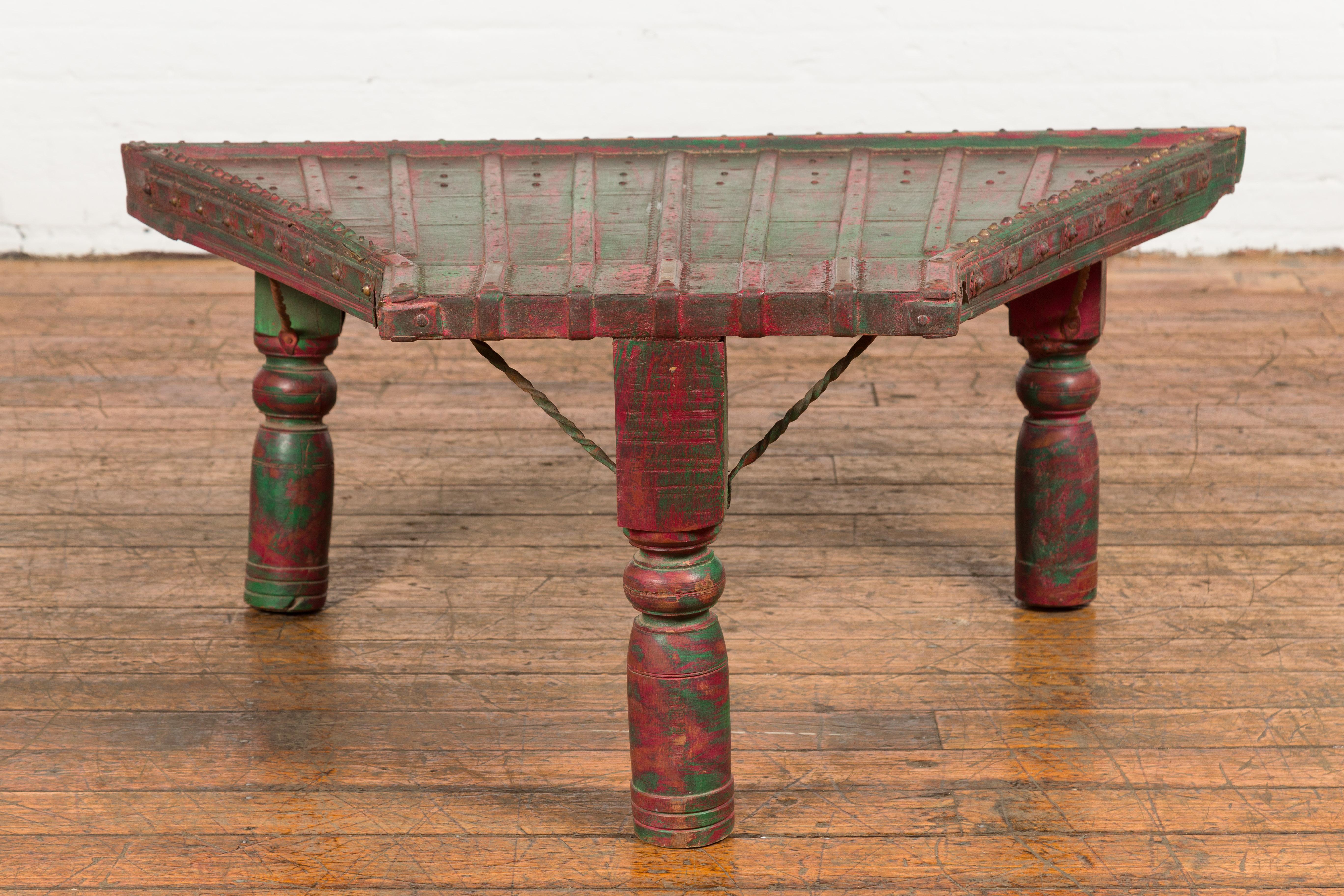 An antique rustic Indian green and red painted coffee table crafted from a repurposed bullock cart from the 19th century with trapezoidal top, protruding front, brass studs, twisted iron stretchers, turned baluster legs and weathered patina.