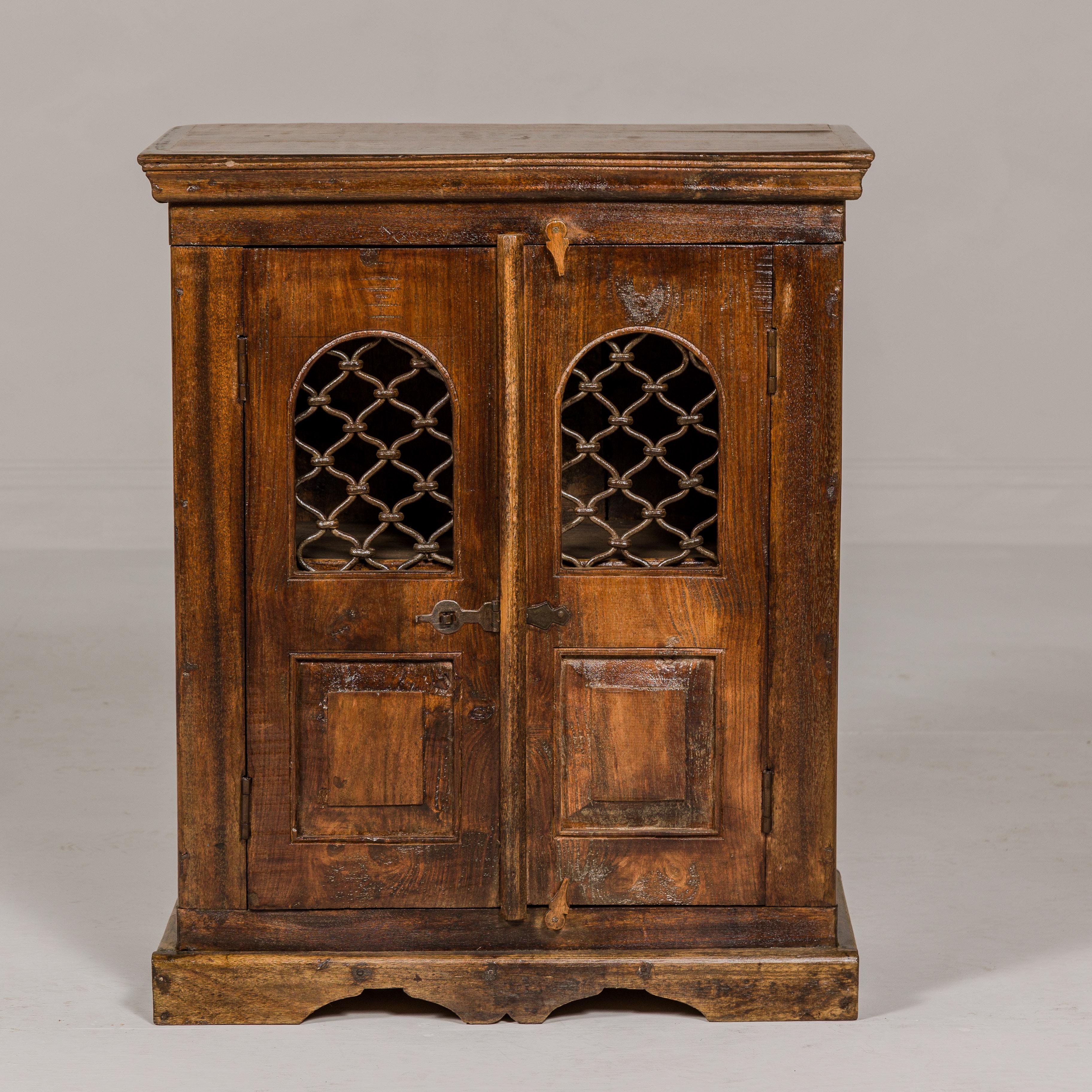 An Indian wooden side cabinet from the 19th century with arched metal grate window door and bracketed plinth. This 19th-century Indian wooden side cabinet is a splendid artifact that marries traditional craftsmanship with functional design. The