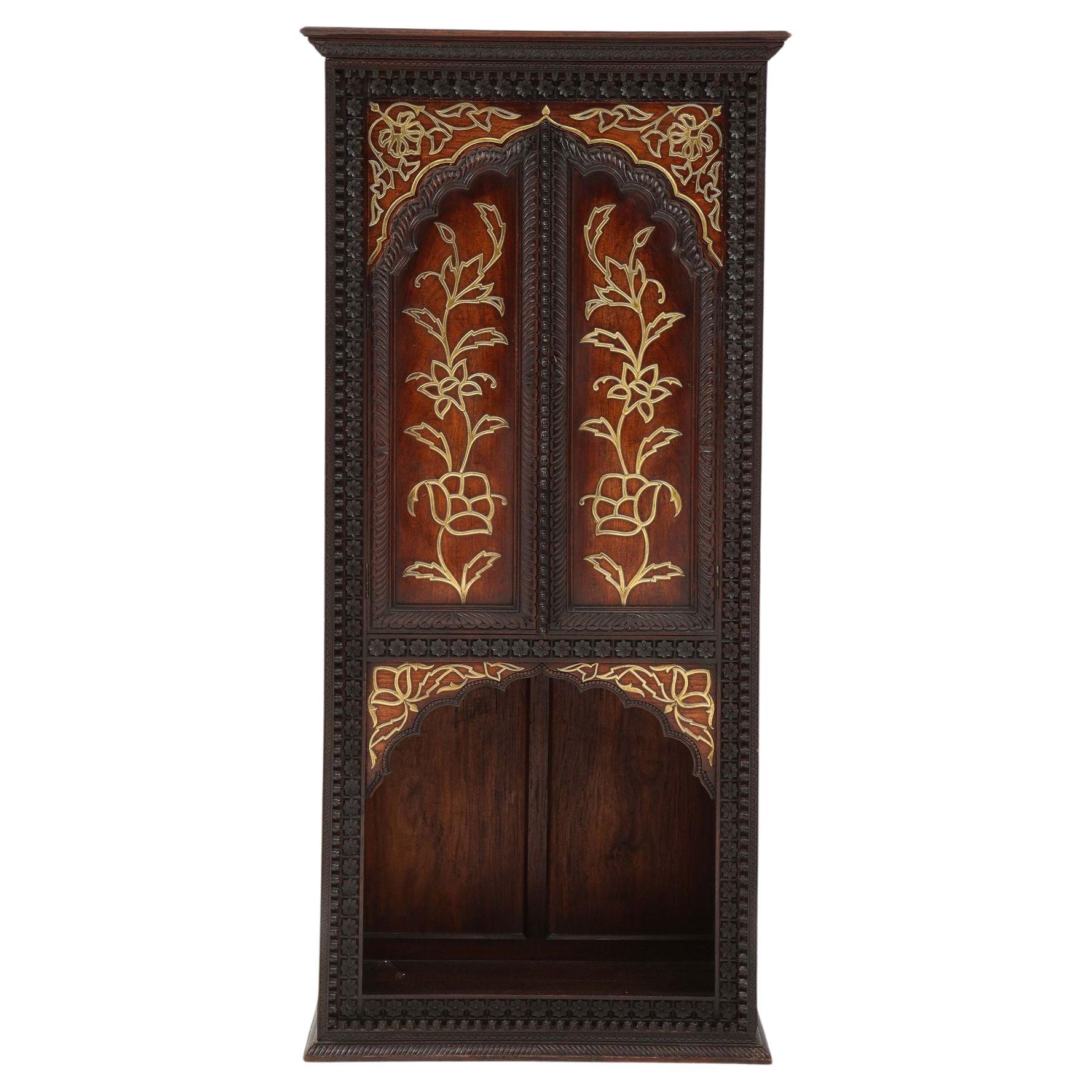 Très belle armoire indienne de la fin du XIXe siècle, en bois dur exotique richement sculpté avec une décoration moulée en laiton dans le goût esthétique, les deux portes arquées au-dessus de la section inférieure ouverte, avec une garniture unique