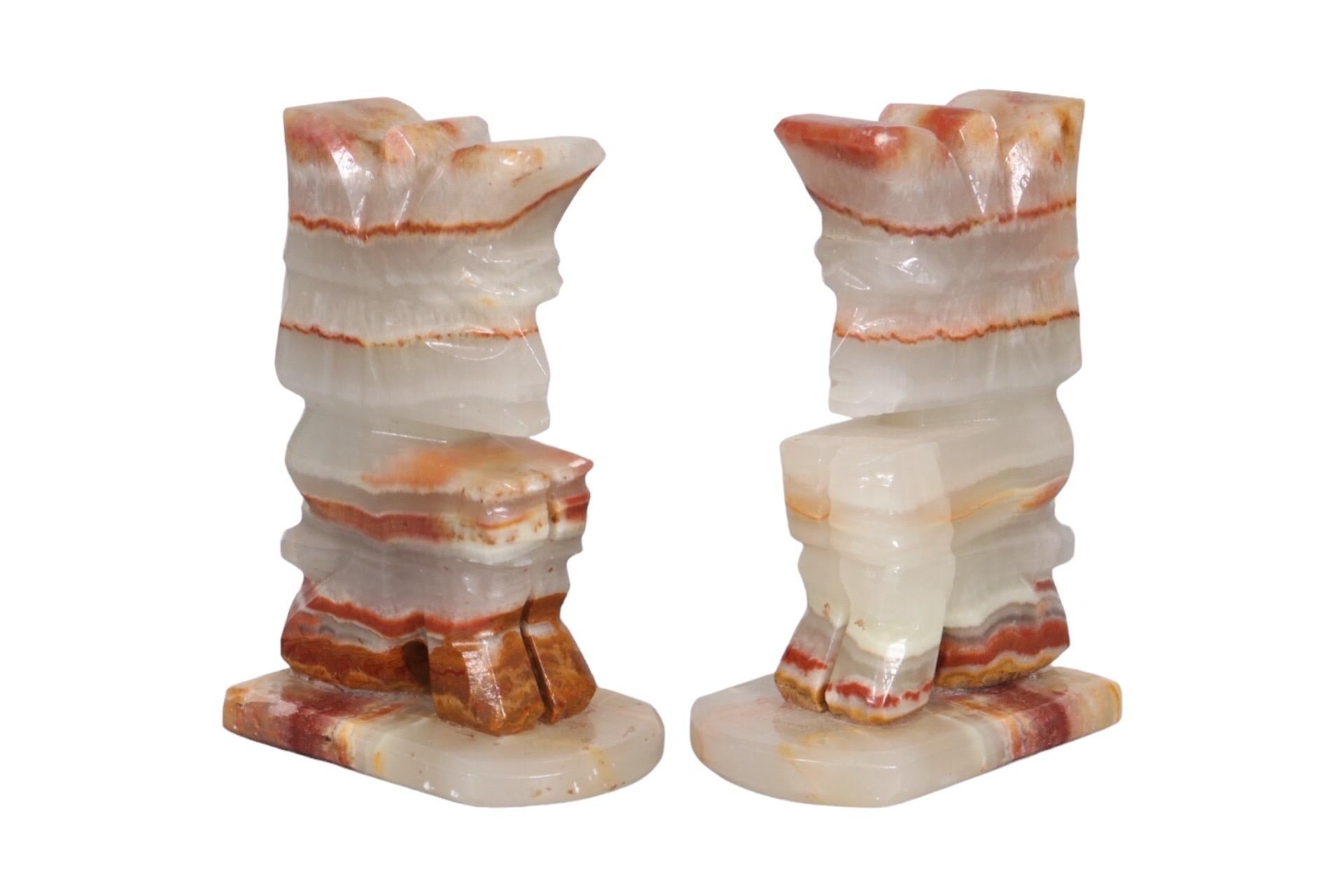 A pair of agate bookends carved in the shape of Indians seated in a hook position. Simple carved lines detail the roach headdress, face and hair. Agate is richly banded in crimson and caramel tones against a translucent off white. Dimensions per