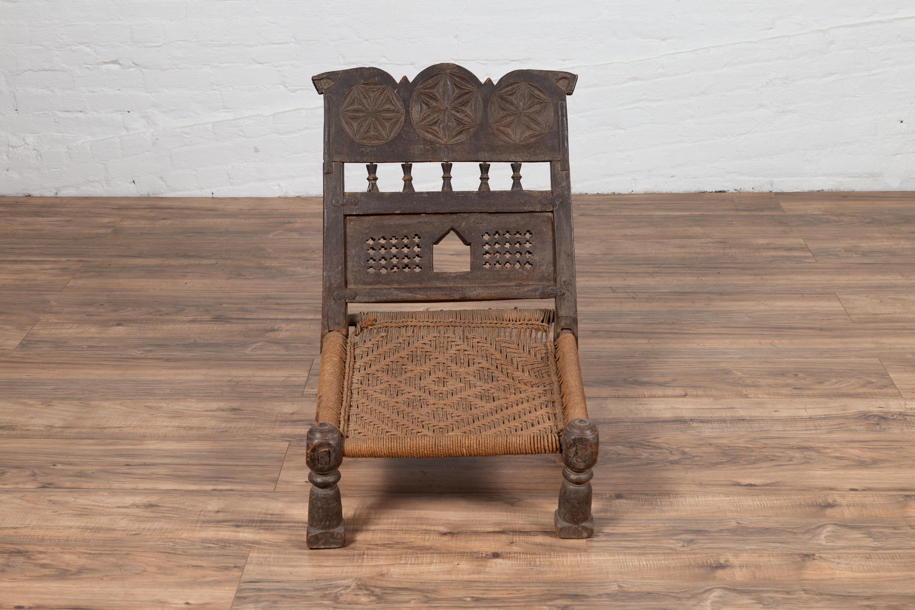 An old Indian rustic wooden low chair from the early 20th century, with handwoven rope seat and carved motifs. Charming our eyes with its rustic appearance, this low seat wooden chair features a short slanted back, adorned with three rosettes hand