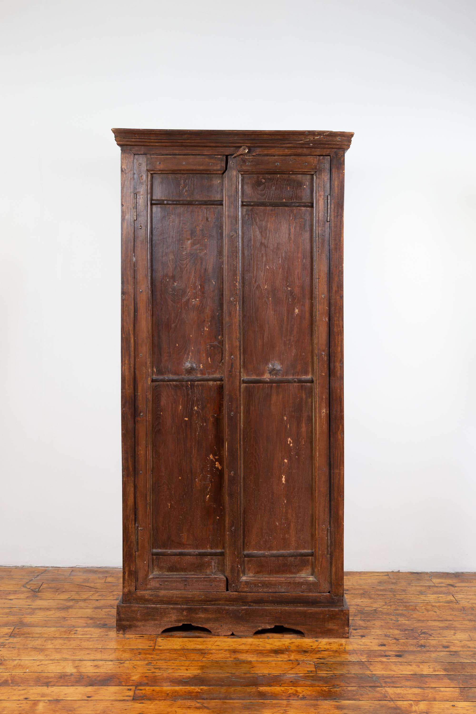 An antique Indian wooden armoire from the early 20th century with paneled doors and interior shelves. Born in India during the early years of the 20th century, this wooden armoire features a linear silhouette, accented with a molded cornice at the