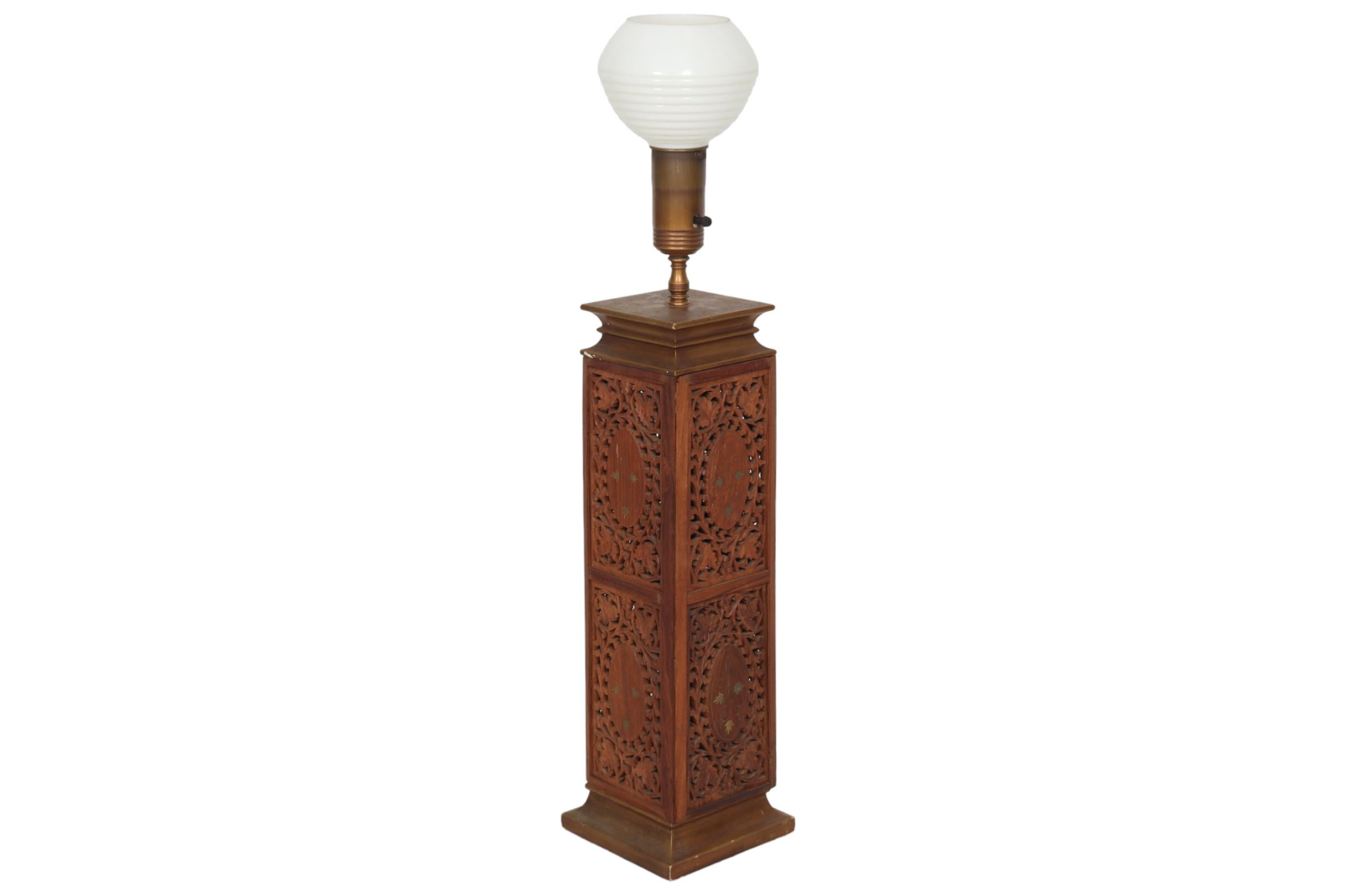 An Indian carved and brass inlaid wooden lamp. An open top ribbed glass globe in white is supported on a highly decorative wooden box. Finely carved and pierced with leaves and berries on a vine. At the center of each face is an oval inlaid with