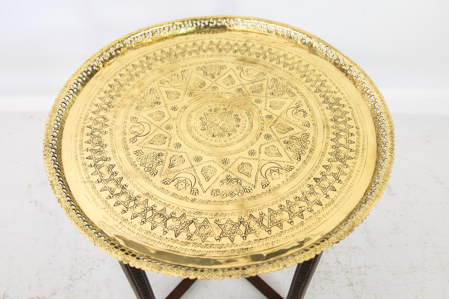 Indian brass top table rests on a carved wooden folding base. The tray is engraved with star and moon motifs, and the gallery is reticulated. The legs are carved on both sides.
 