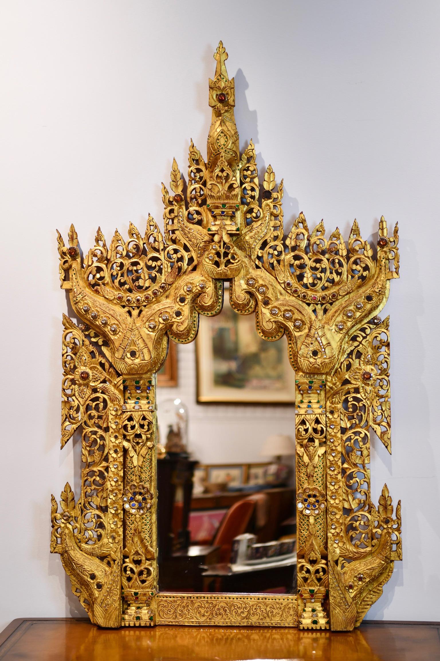 Ornate heavily carved and giltwood mirror. The wood is gilded throughout and adorned with colorful glass pieces. Some glass pieces are missing but overall very good condition, including mirror glass. Likely Thai or Burmese. Dimensions: 56