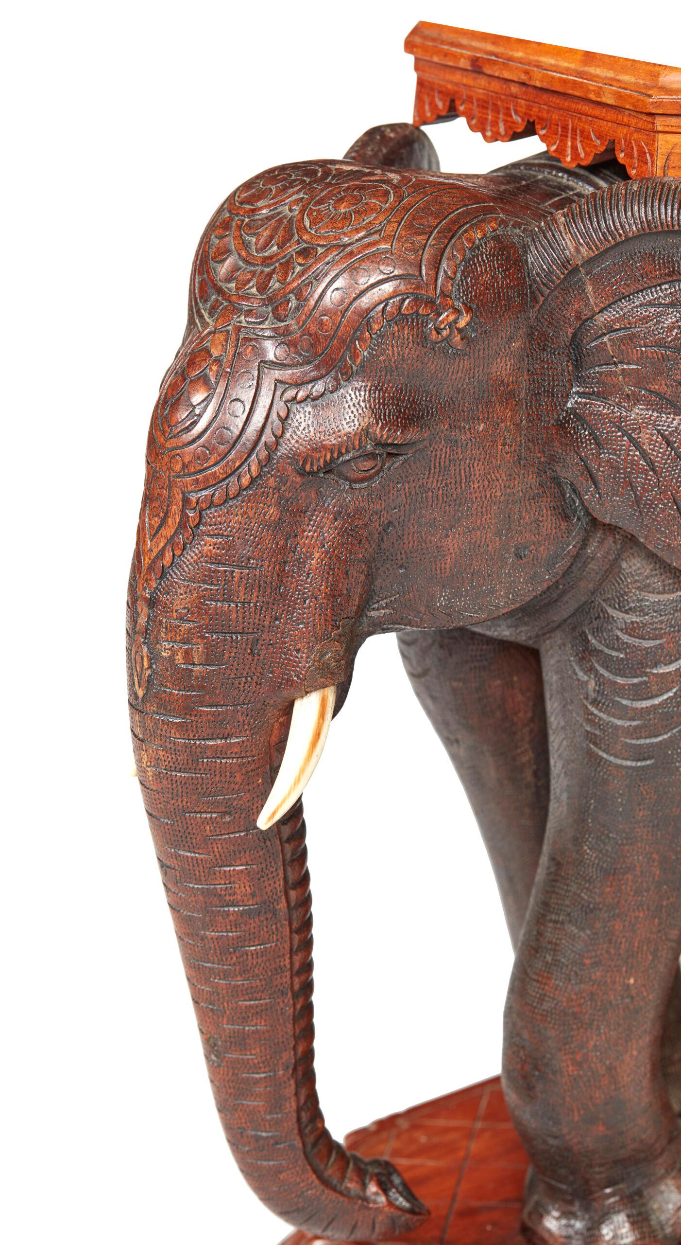 Mesa de elefante tallada en madera dura de la India, La tapa octogonal con un delantal festoneado descansa sobre el lomo de un elefante montado con colmillos de marfil, sobre una base oblonga tallada con puntas de hojas y galones, con la inscripción