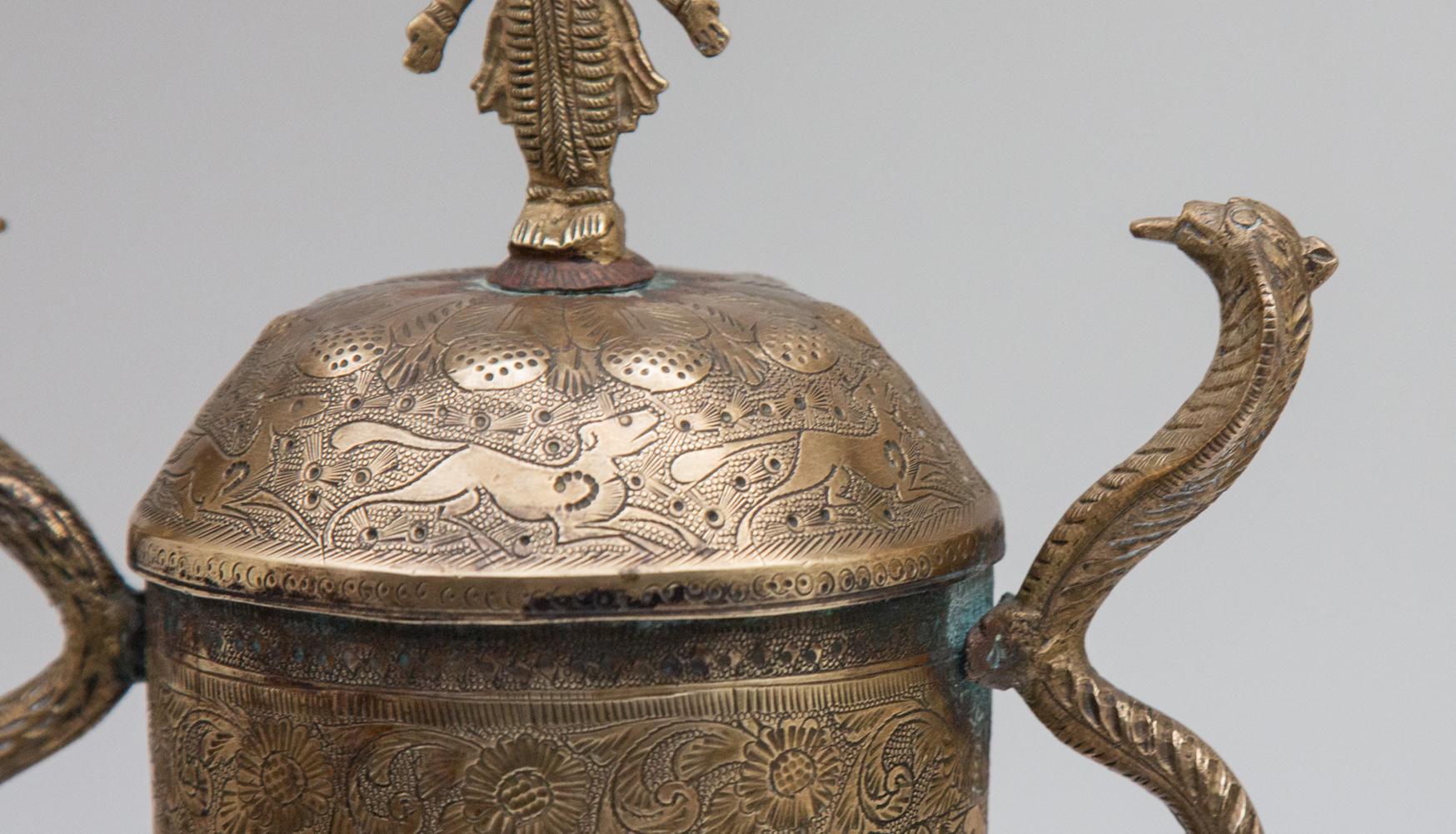 Indian covered brass urn with deity and serpent handles. Two handles made of cobras, engraved with dogs, flowers, and foliage. Topped with four-armed deity. Measures: 13