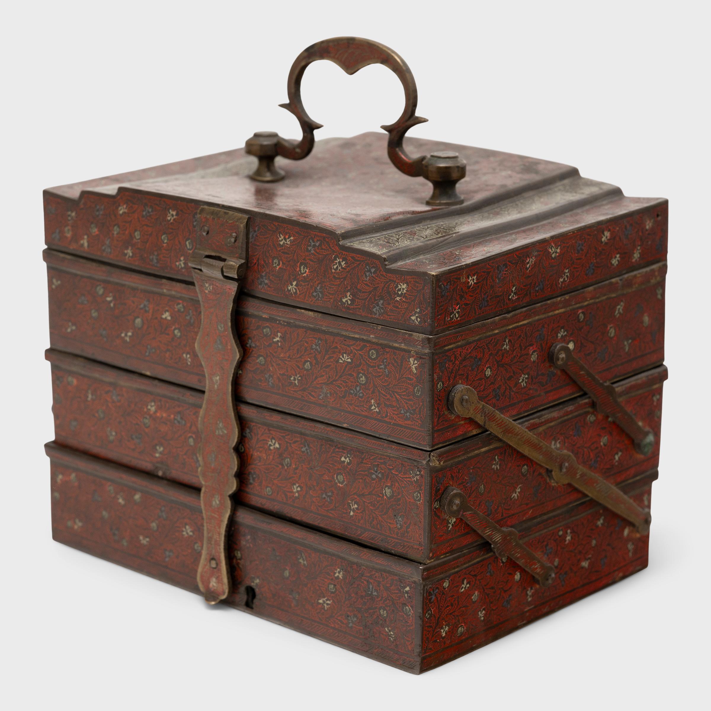 Cette boîte indienne à trois niveaux a été fabriquée au début du XXe siècle et était à l'origine utilisée comme coffre de toilette ou boîte à bijoux. Gravée d'un élégant motif floral, la boîte en laiton est recouverte d'un riche pigment rouge aux