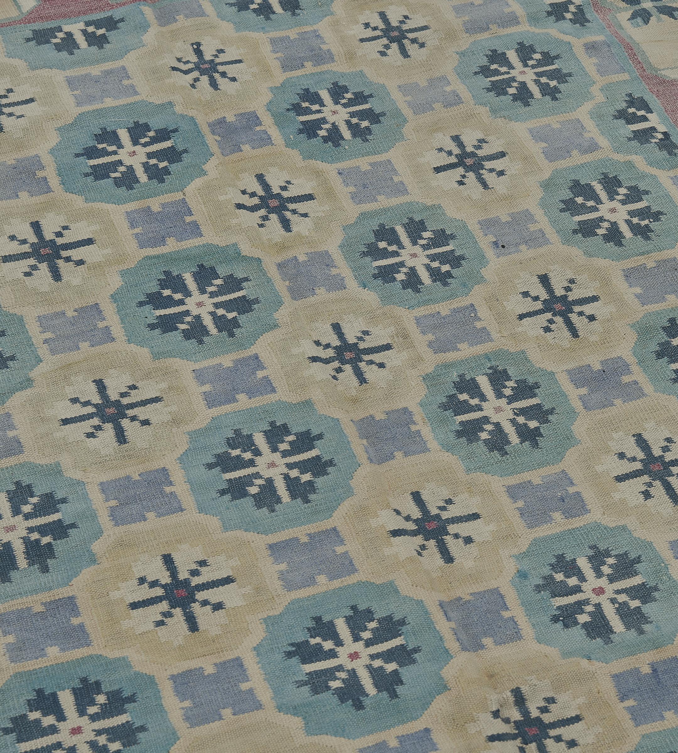 This hand-woven wool antique mid-20th century Indian Dhurrie rug features a geometric pattern with repeating floral-like motifs in the central field, arranged in a grid layout. The primary color palette consists of soft pastel shades, including