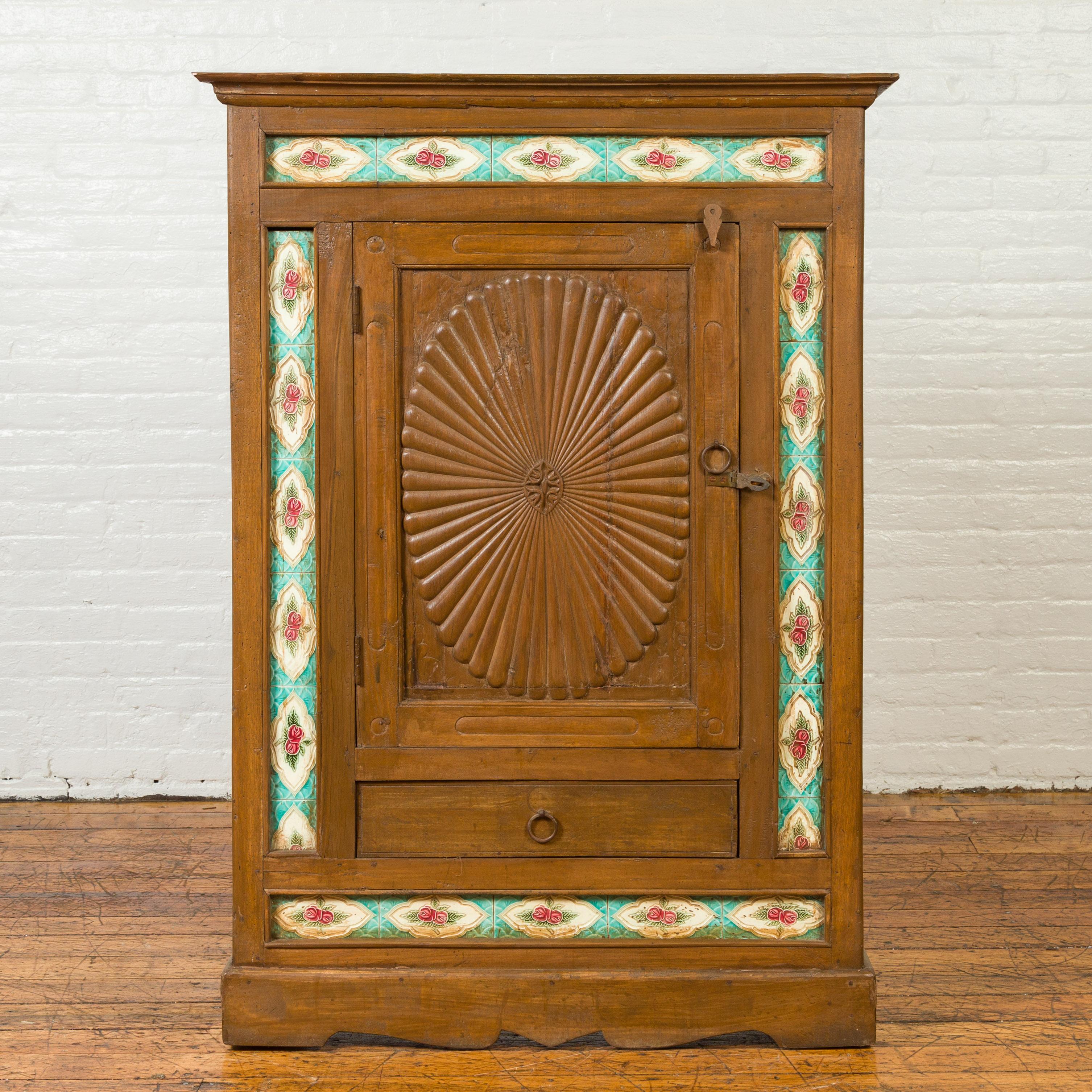 An antique Indian small cabinet from the early 20th century with carved sunburst pattern and hand painted tiles. Born in India, this wooden cabinet features a small cornice overhanging a perfectly organized façade. A central door, carved with oval