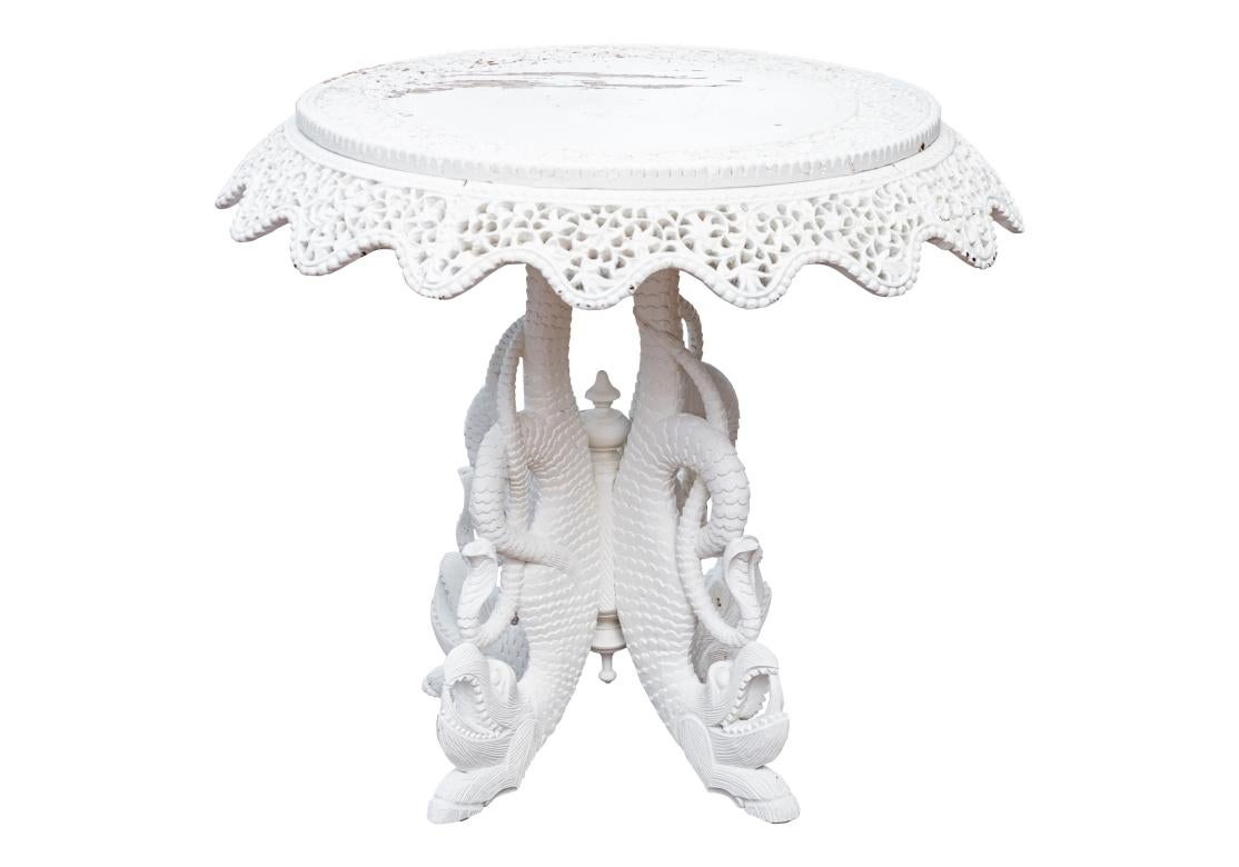 An elaborately carved and white painted India occasional pedestal table. The circular table top with carved foliate scrolled and beaded bands above the carved, pierced and scalloped foliate scrolled apron. Mounted on an interesting base with four