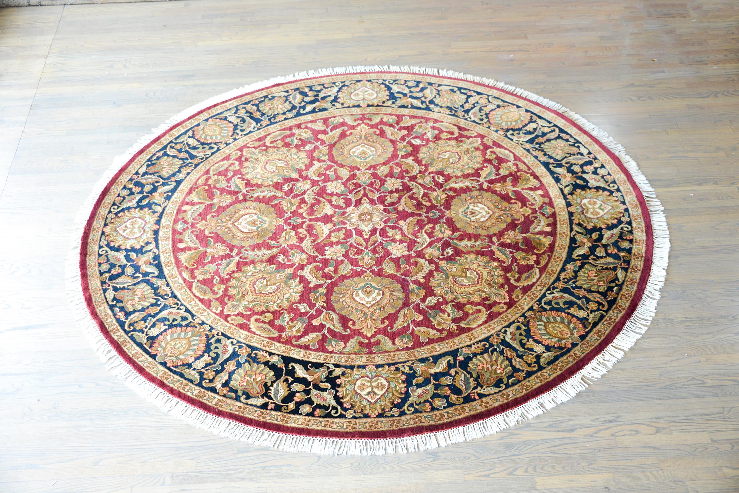 A beautiful vintage Indian Tabriz-style round rug with a mirrored pattern in the center with several stylized flowers and scrolling vines, woven in gold, cream, orange, and white, against a cranberry background. The border is similarly patterned