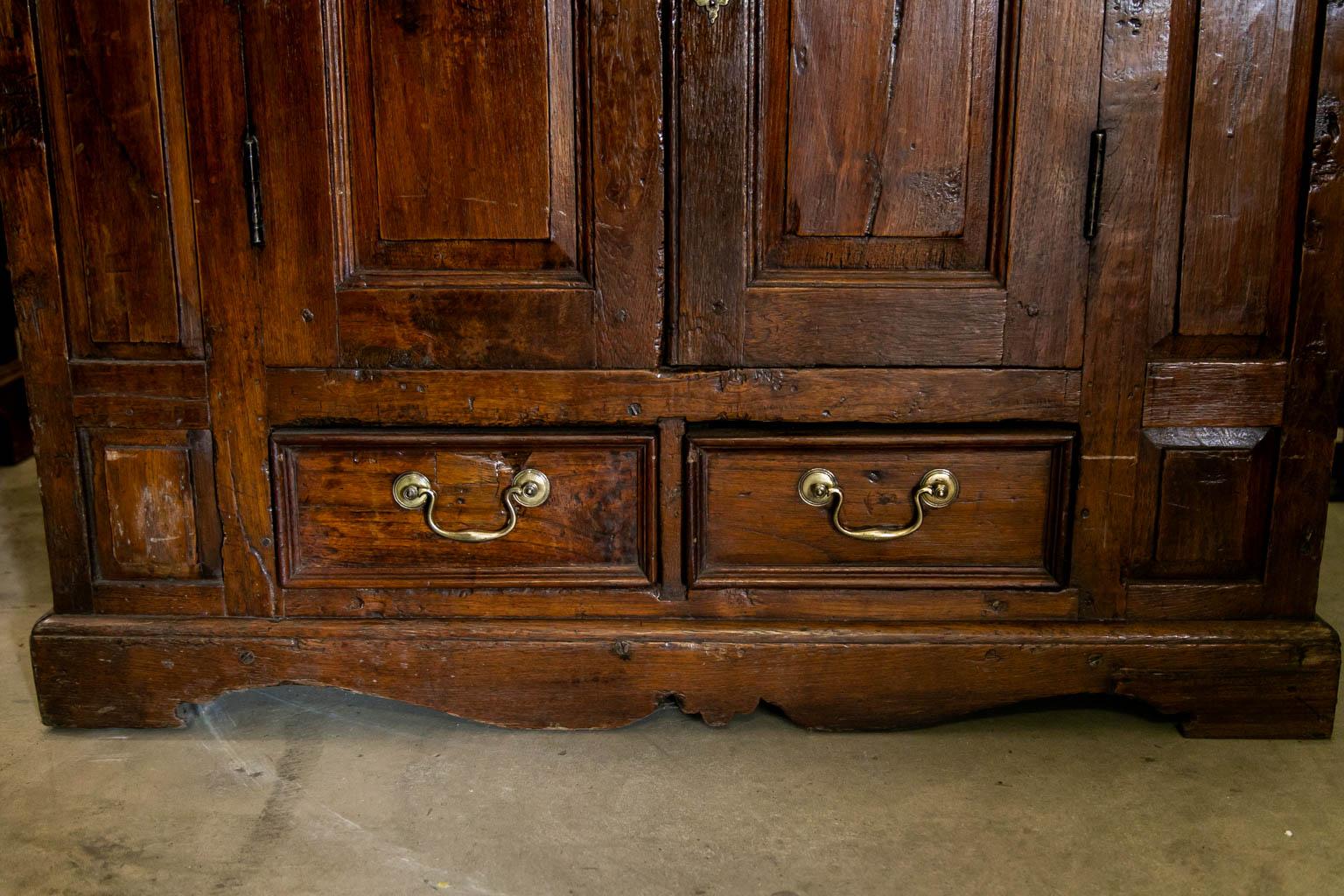 This four door/two drawer cupboard has exposed peg construction. There are raised panels on the doors and stiles. The hinges are original and brass hardware is later. The top section has one fixed shelf. The lower section is open.