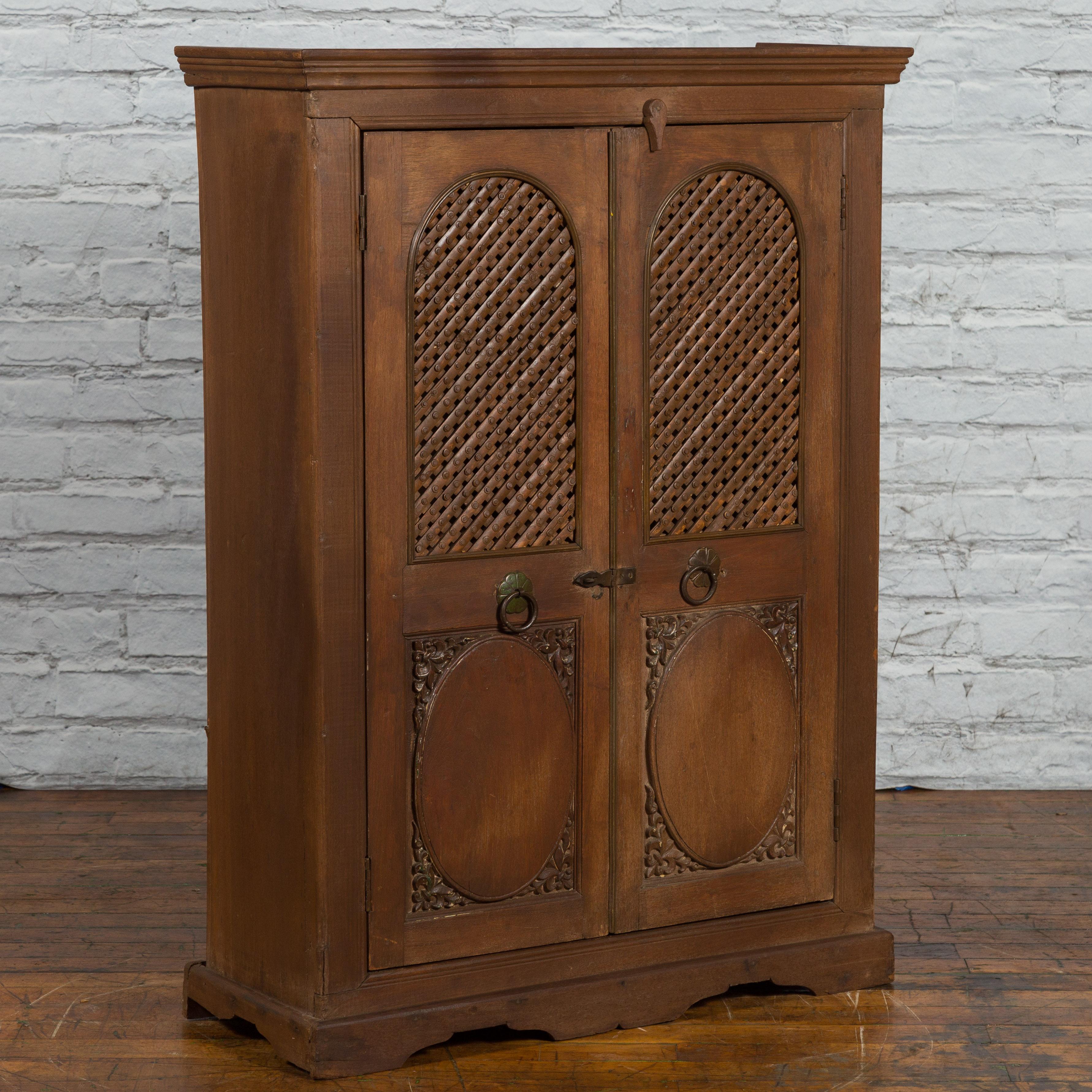 An Indian wooden cabinet from the mid 20th century, with lattice motifs and carved panels. Created in India during the midcentury period, this wooden cabinet features a molded cornice sitting above two doors adorned with arching panels and lattice