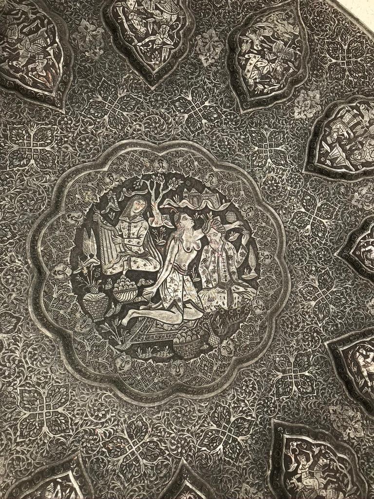 A stunning embossed tray with a central erotic scene of a turbaned male and a topless woman on a Persian carpet with a harp player, bowls of fruit, a carafe of wine and vase with flowers. Surrounded by 12 embossed cartouches with various