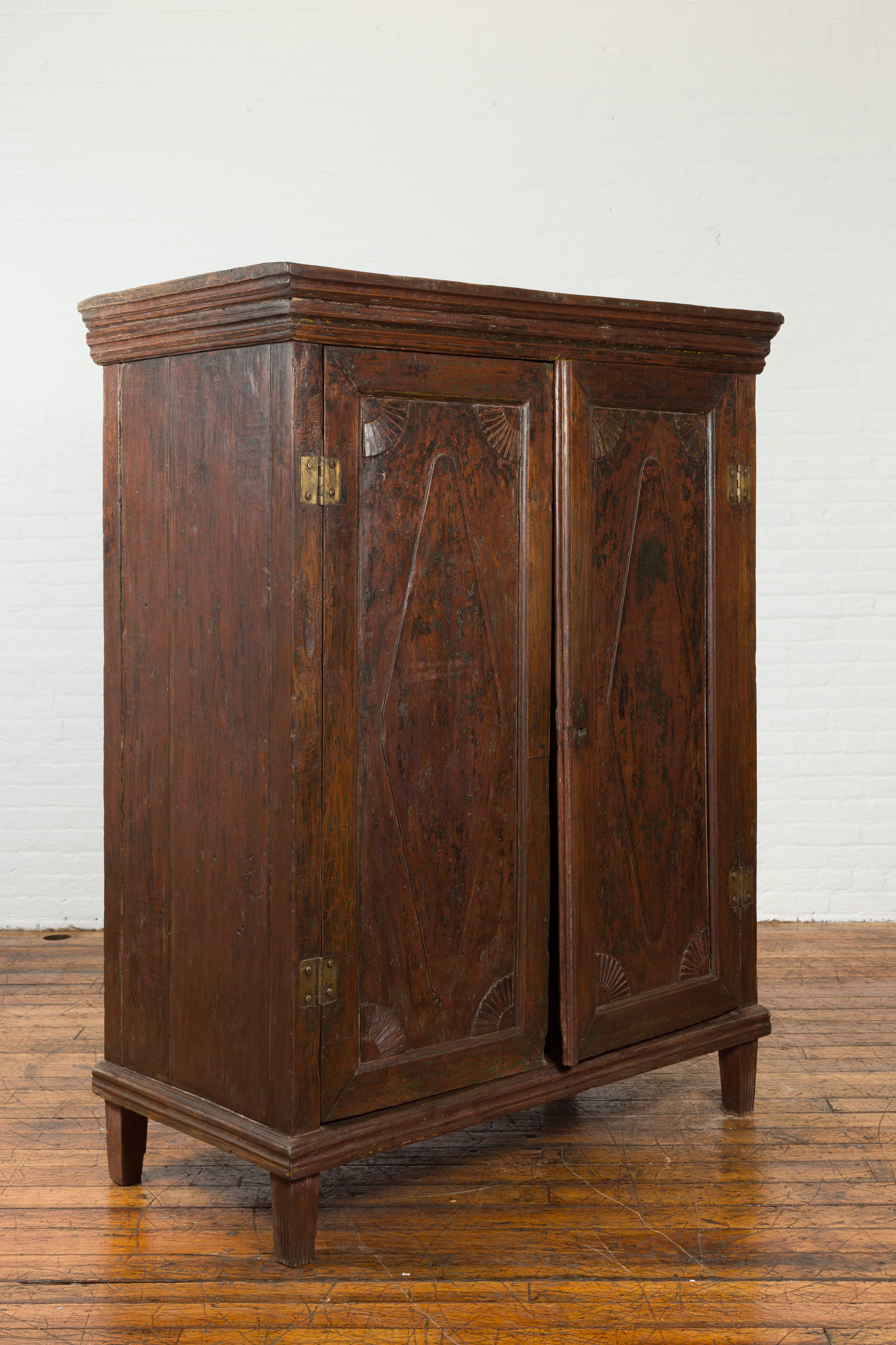 An Indonesian large antique cabinet from the 19th century with elongated diamond shapes on the front of the double doors and scalloped fans featured on each of the door's corners. Our tall teak wood antique cabinet features a molded bordered top