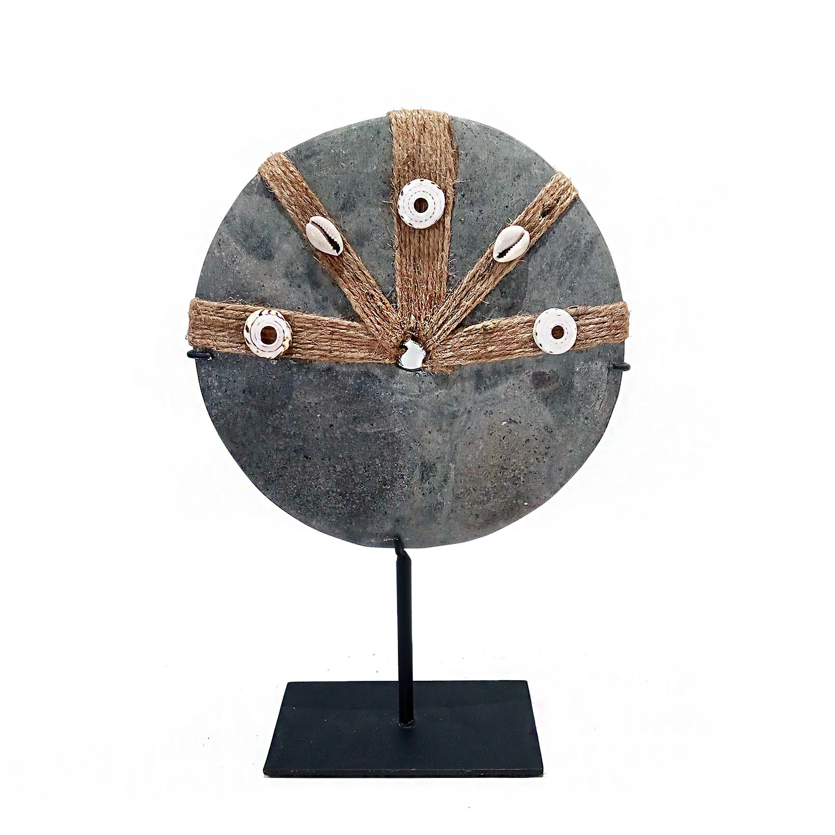 A decorative stone coin from Bali, Indonesia. Grey stone with raffia rope and seashell appliques. Mounted on a made-to-measure black metal stand. 

 8 inches in diameter, 12 inches high, on a black metal stand with a 3