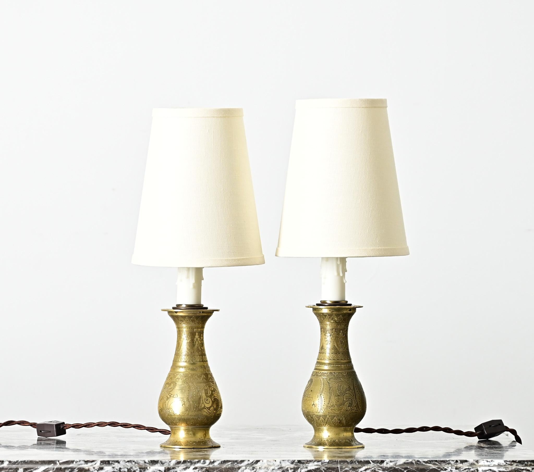 A pair of 20th century petite brass vases or candlesticks with Indonesian design motifs, that were adapted and electrified as custom table lamps. Their history is particularly evident in the depictions of traditional shadow puppet figures found