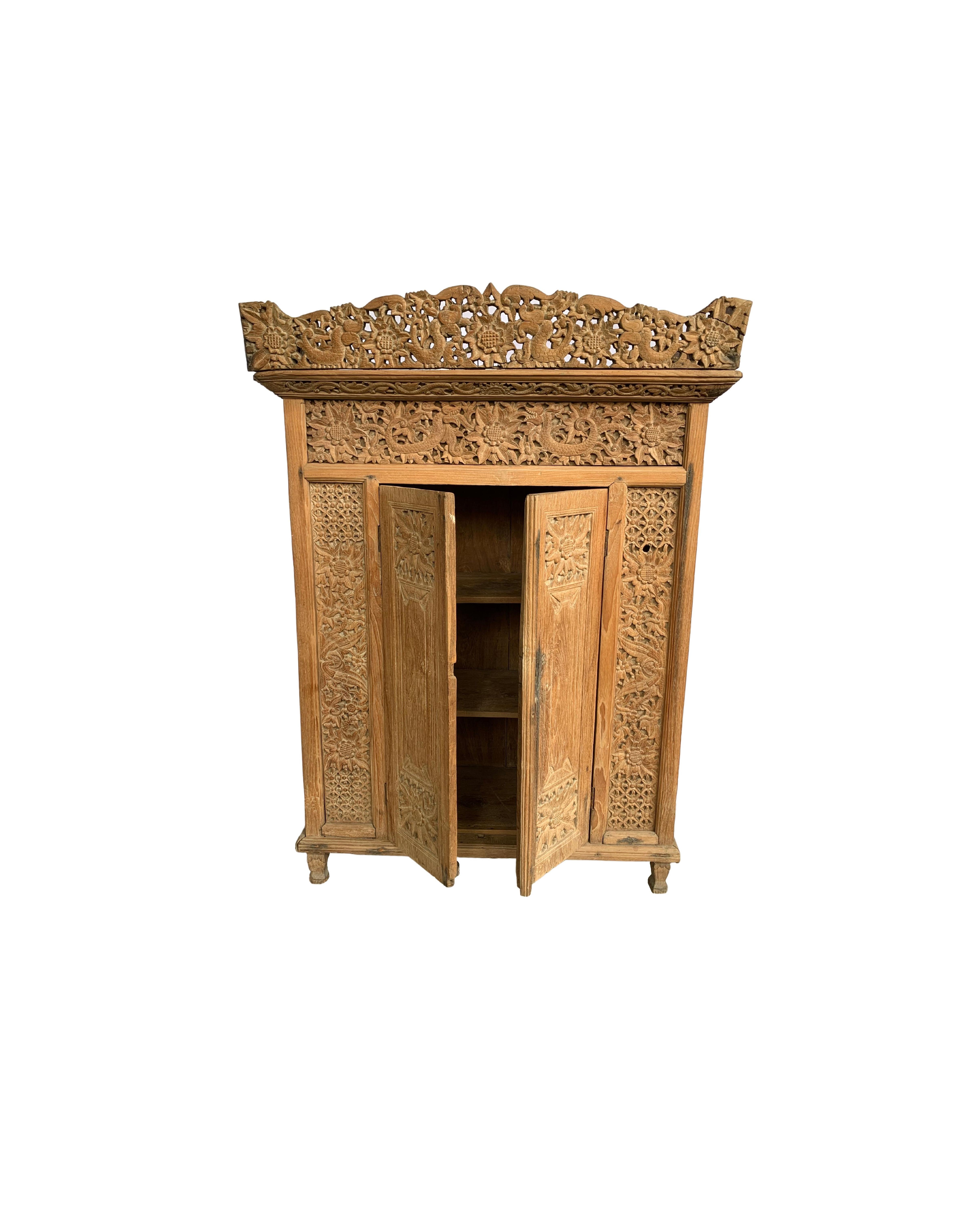 This teak cabinet dates to the Mid-20th Century and features an elaborate exterior largely covered by hand-carved motifs. The motifs are largely floral but also features scaly mythical creatures. The front doors open to 3 interior shelves that