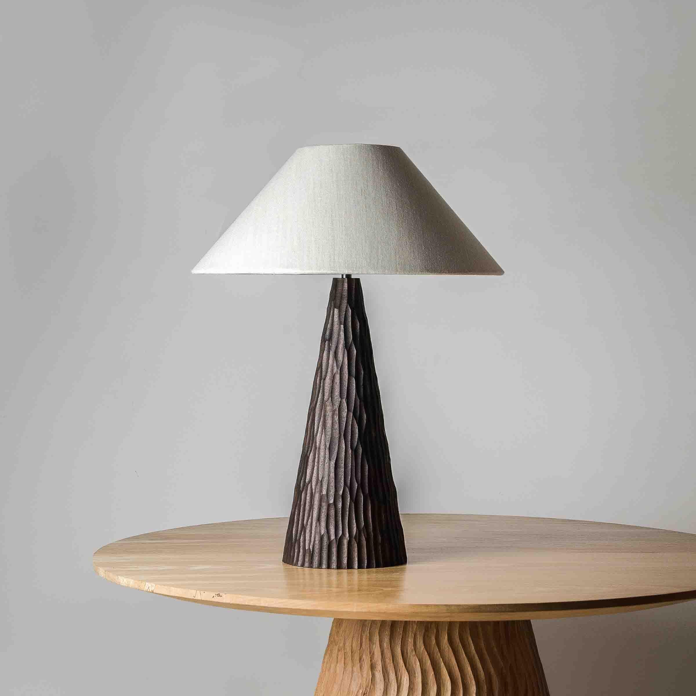 The Indra table lamp - black pairs a sculptural wooden base with a steeply angled lampshade, that diffuses light downwards to create a cosy setting.

The conical base is turned and carved by hand from solid ash, and stained black.

The shade is