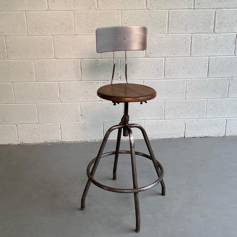 Industrial, drafting stool features a brushed steel frame and back with a 14 inch round, swivel, maple seat that is height adjustable from 27.5 - 31 inches.
