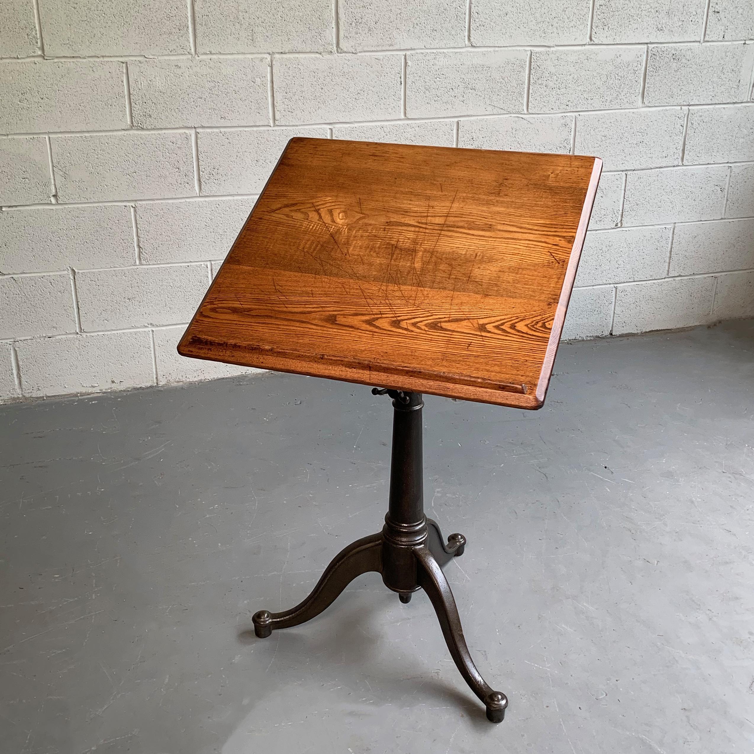 Small, antique, artist rendering easel or drafting table features an oak top with tool ledge on a tilt and height adjustable, dark, cast iron, pedestal base that adjusts from 29 - 40 inches height. The table features engineer supply company, Kolesch