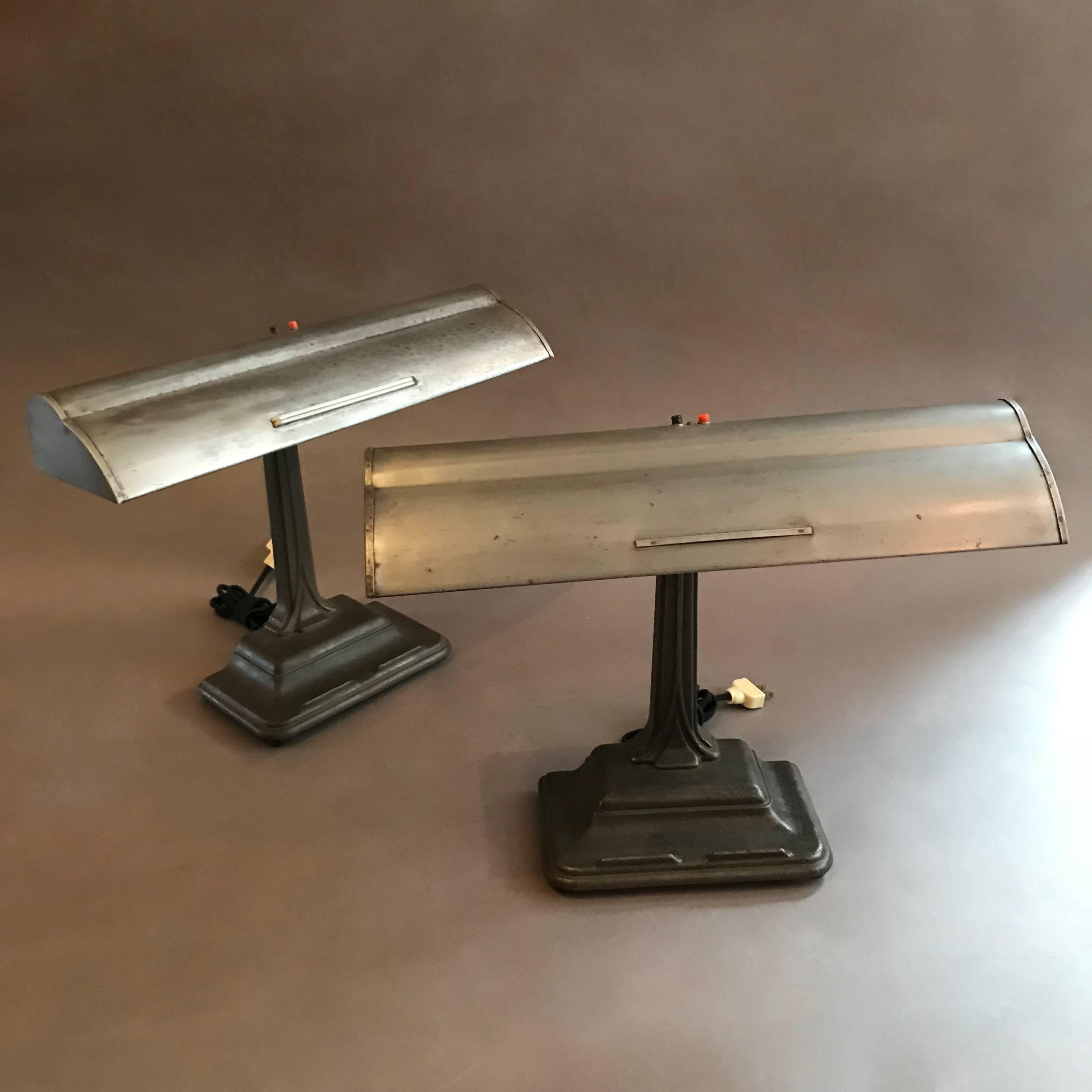 Classic, 1930s, banker's desk lamp features a cast iron base and neck with brushed steel shade. The lamps each have a porcelain medium socket that accepts two bulbs. Two available. Base measures: 9in. W x 6in. D. Please note that the metal grid