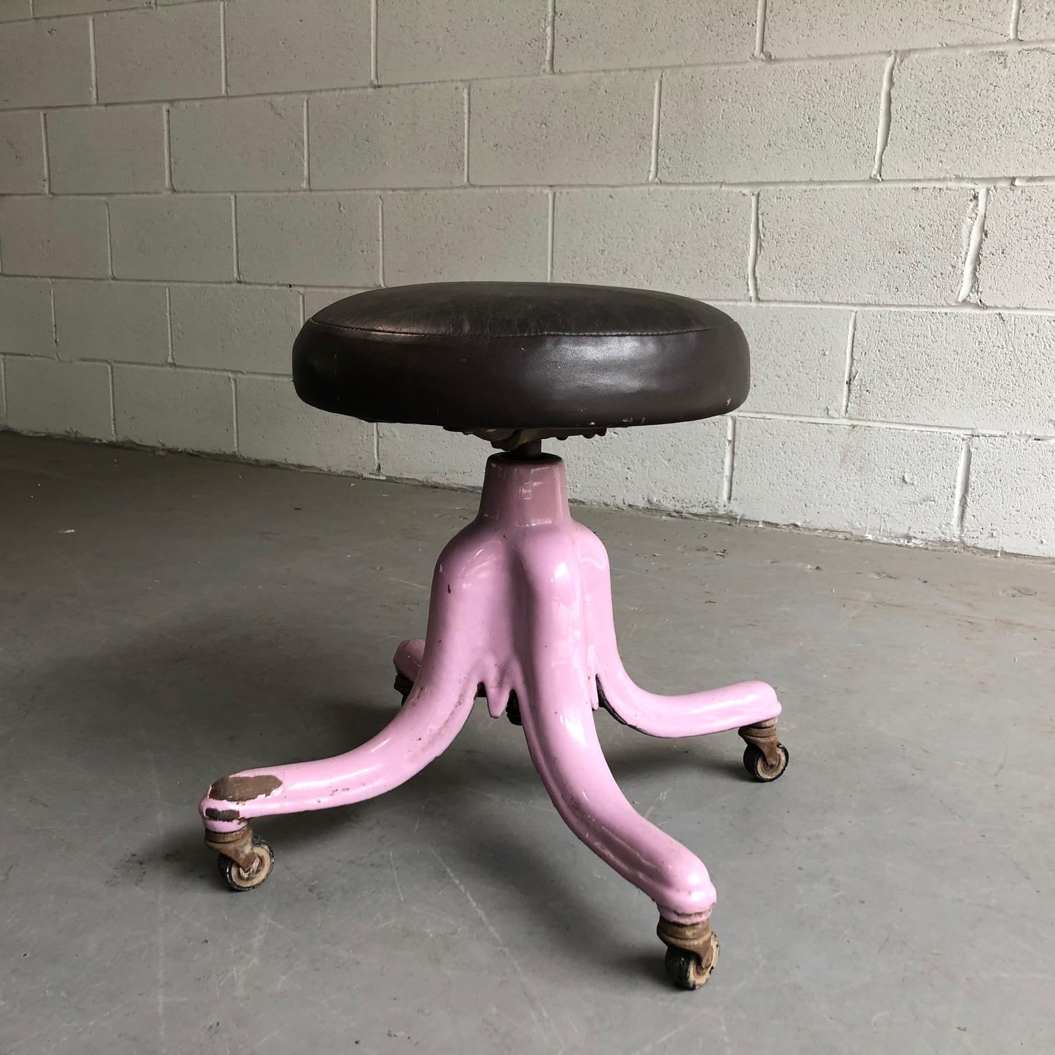 Industrial, rolling, medical stool features a pink hued, baked enamel steel frame with 13.5 inch diameter, dark brown leather seat that is height adjustable from 18-25”.