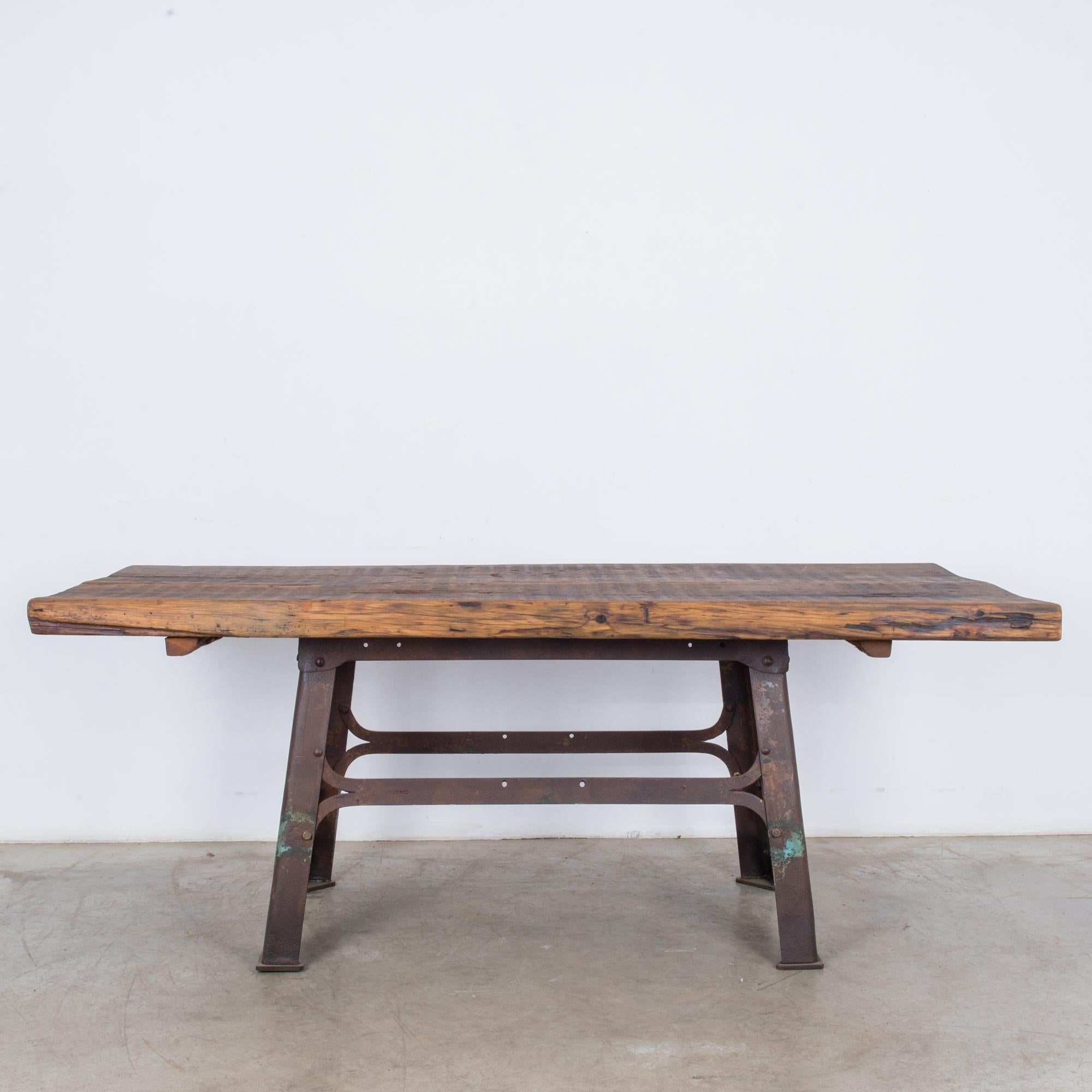 A table from Belgium, circa 1900, with a metal base and wooden top. Legs of metal girders, set at an A-frame angle, make an industrial contrast with the organic swell of the tabletop; a rustic wooden slab, marked by soft undulation and a dark grain.