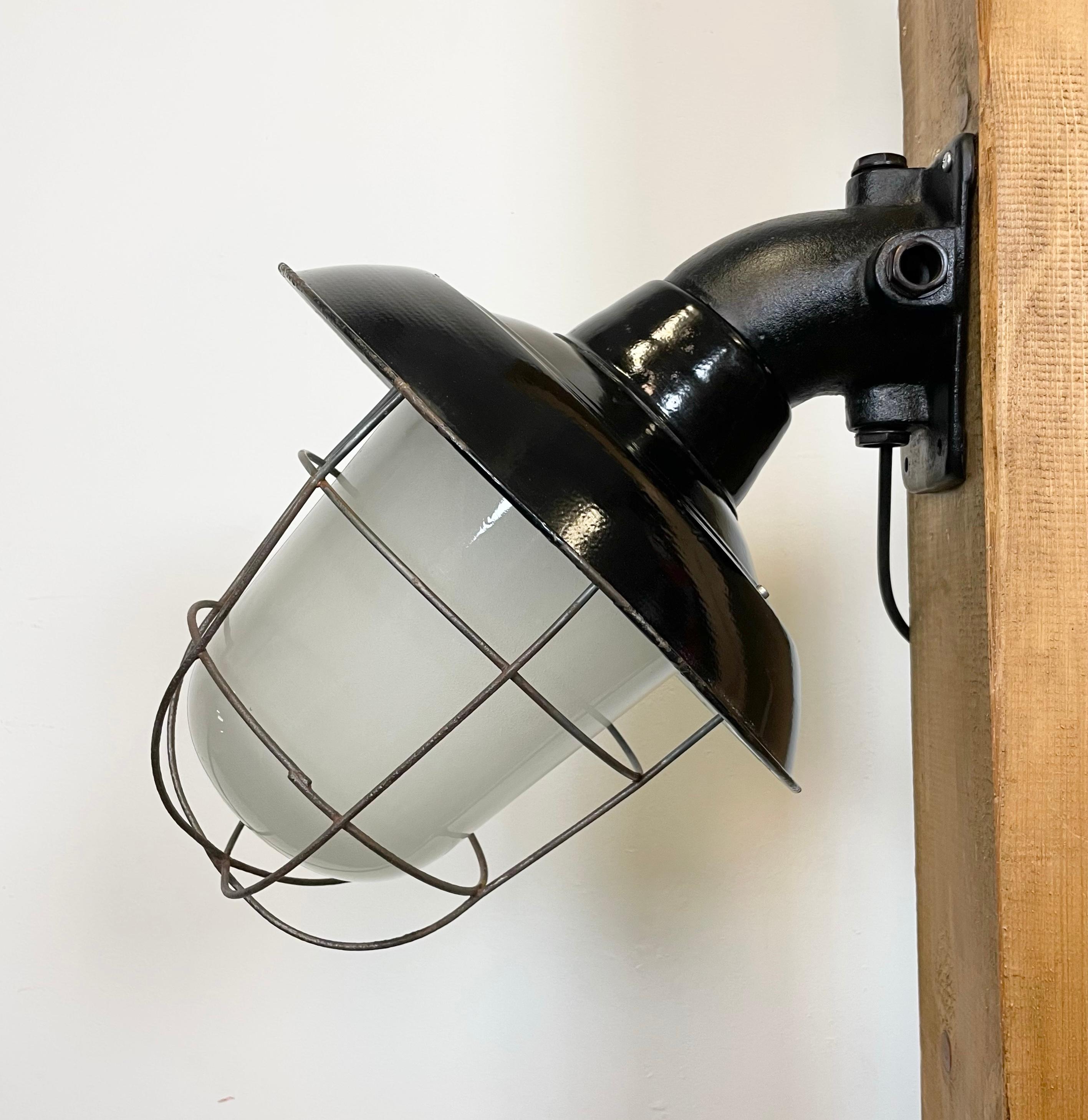 - Industrial wall lamp with cast iron wall mouting from former Czechoslovakia made during the 1960s
- Black enamel shade with white enamel interior, milk glass, iron grid
- New porcelain socket for E 27 light bulbs and wire
- Weight: 5 kg.