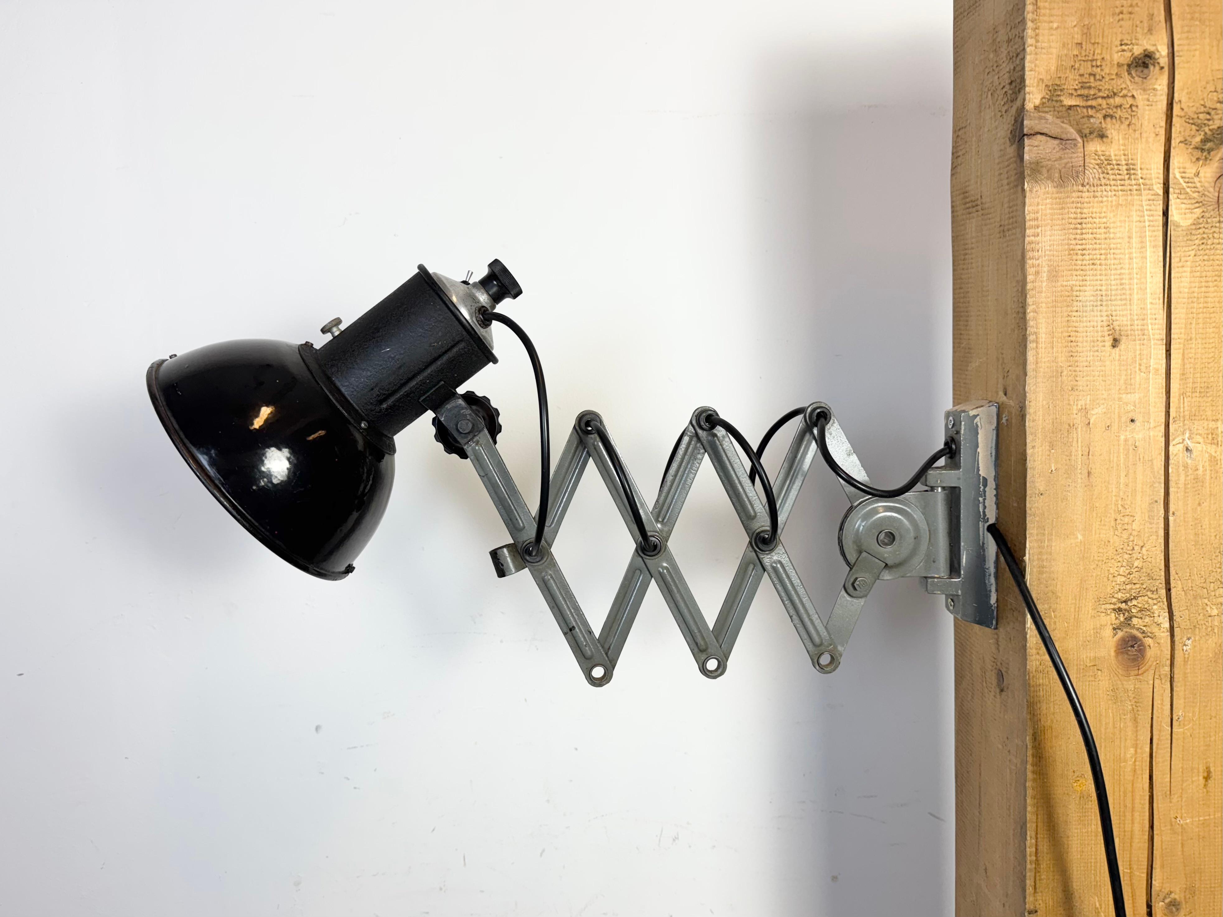 Cette lampe murale à ciseaux industrielle ajustable vintage a été produite dans l'ancienne Tchécoslovaquie au cours des années 1950. La lampe a un abat-jour en métal émaillé noir avec intérieur émaillé blanc. Le bras de ciseaux en fer gris est