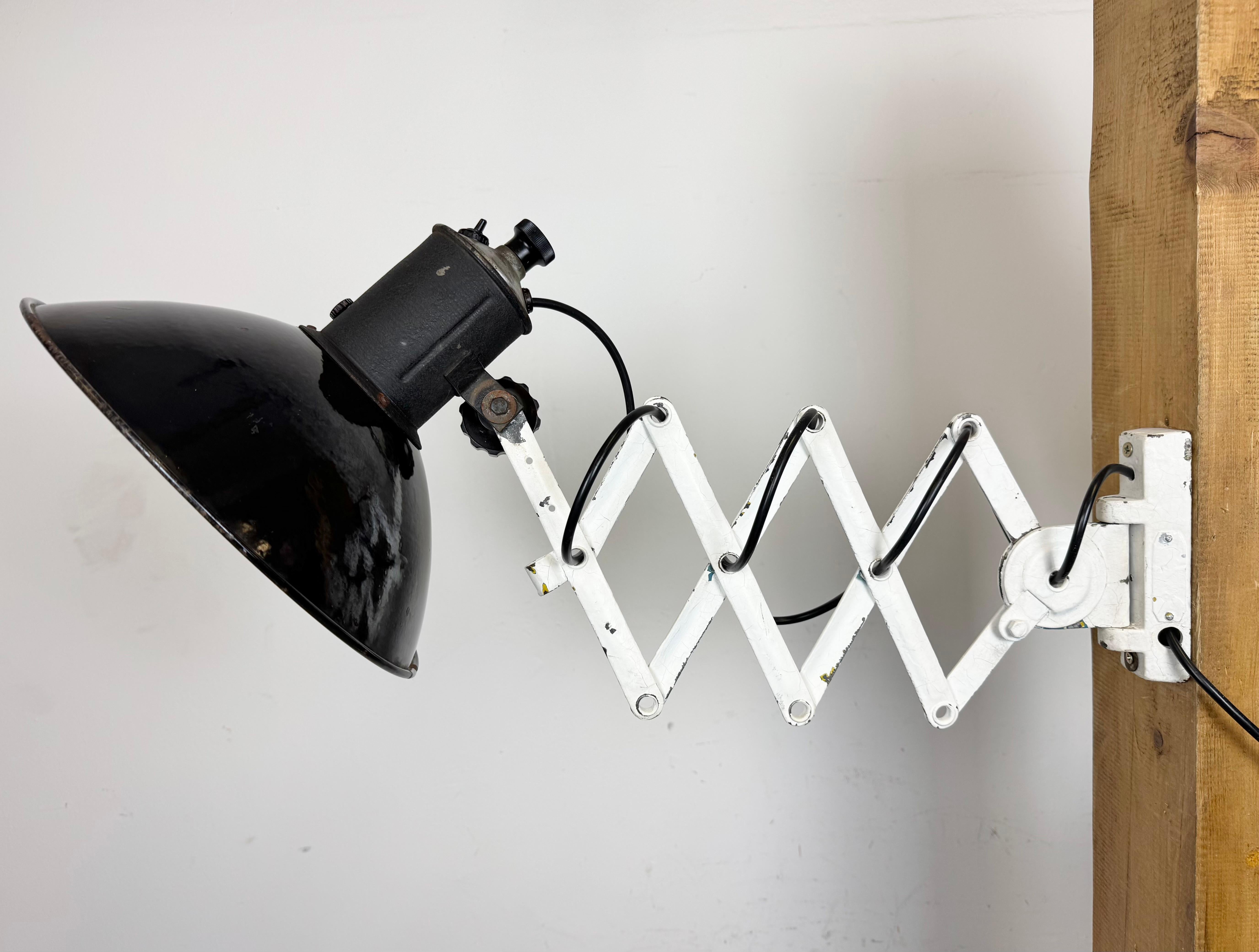 This vintage industrial adjustable scissor wall lamp was produced in former Czechoslovakia during the 1950s. The lamp has a black enamel metal shade with white enamel interior. White iron scissor arm is extendable and can be turned sideways.