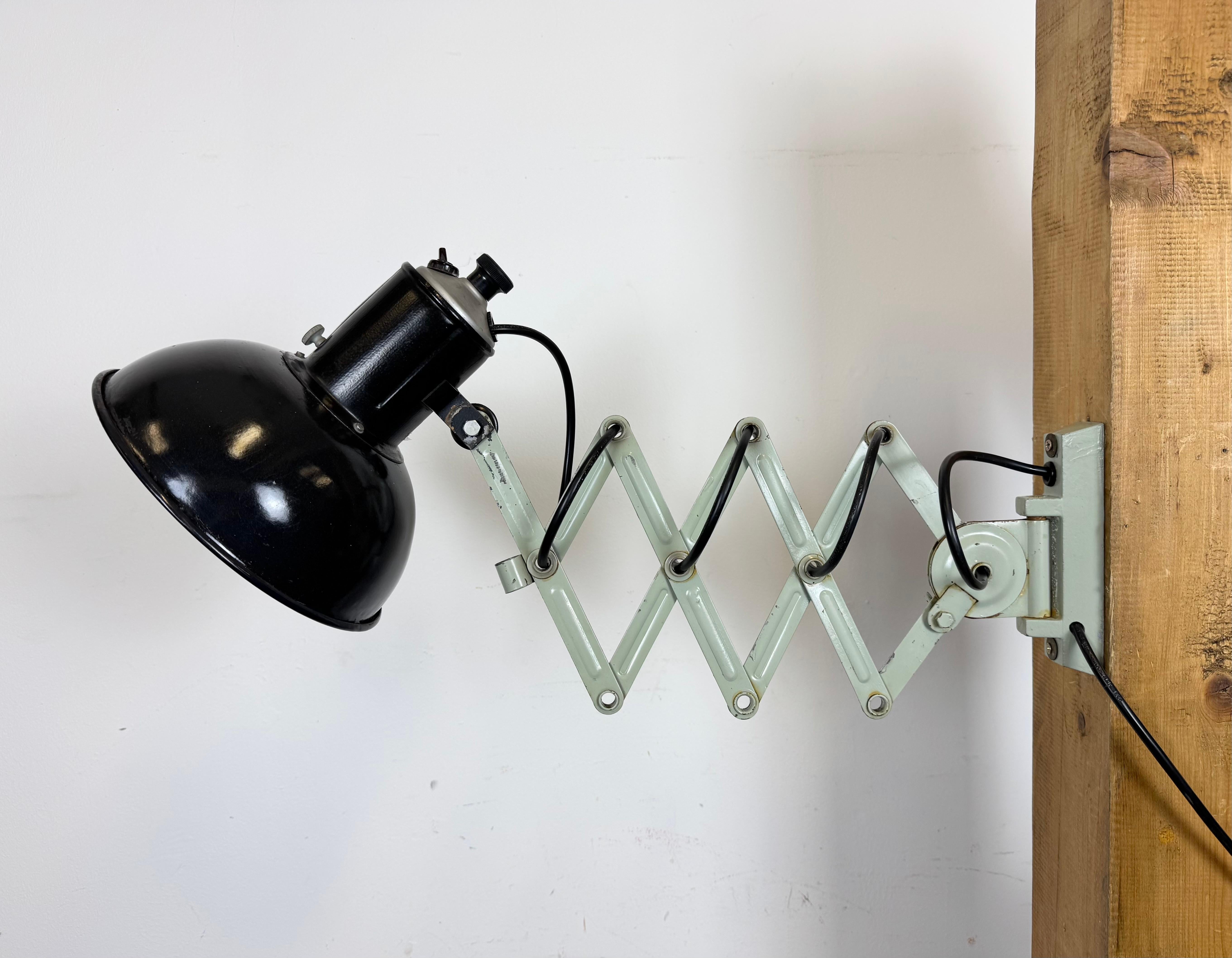 Cette applique industrielle vintage à ciseaux ajustables a été produite dans l'ancienne Tchécoslovaquie au cours des années 1950. La lampe a un abat-jour en métal émaillé noir avec intérieur émaillé blanc. Le bras de ciseaux en fer léger geeen est