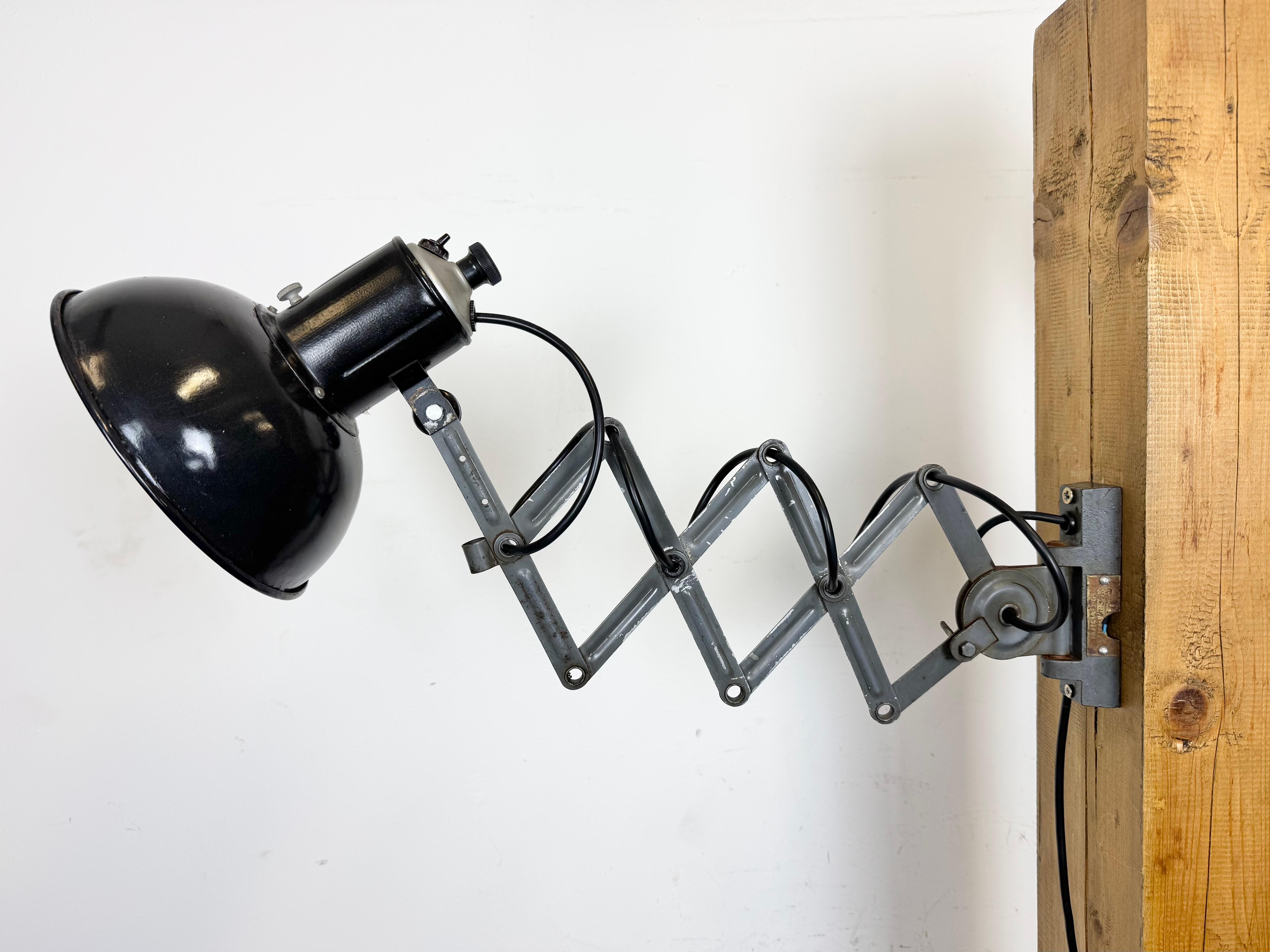 Cette lampe murale à ciseaux ajustable de style industriel vintage a été produite dans l'ancienne Tchécoslovaquie au cours des années 1950. La lampe a un abat-jour en métal émaillé noir avec intérieur émaillé blanc. Le bras de ciseaux en fer gris