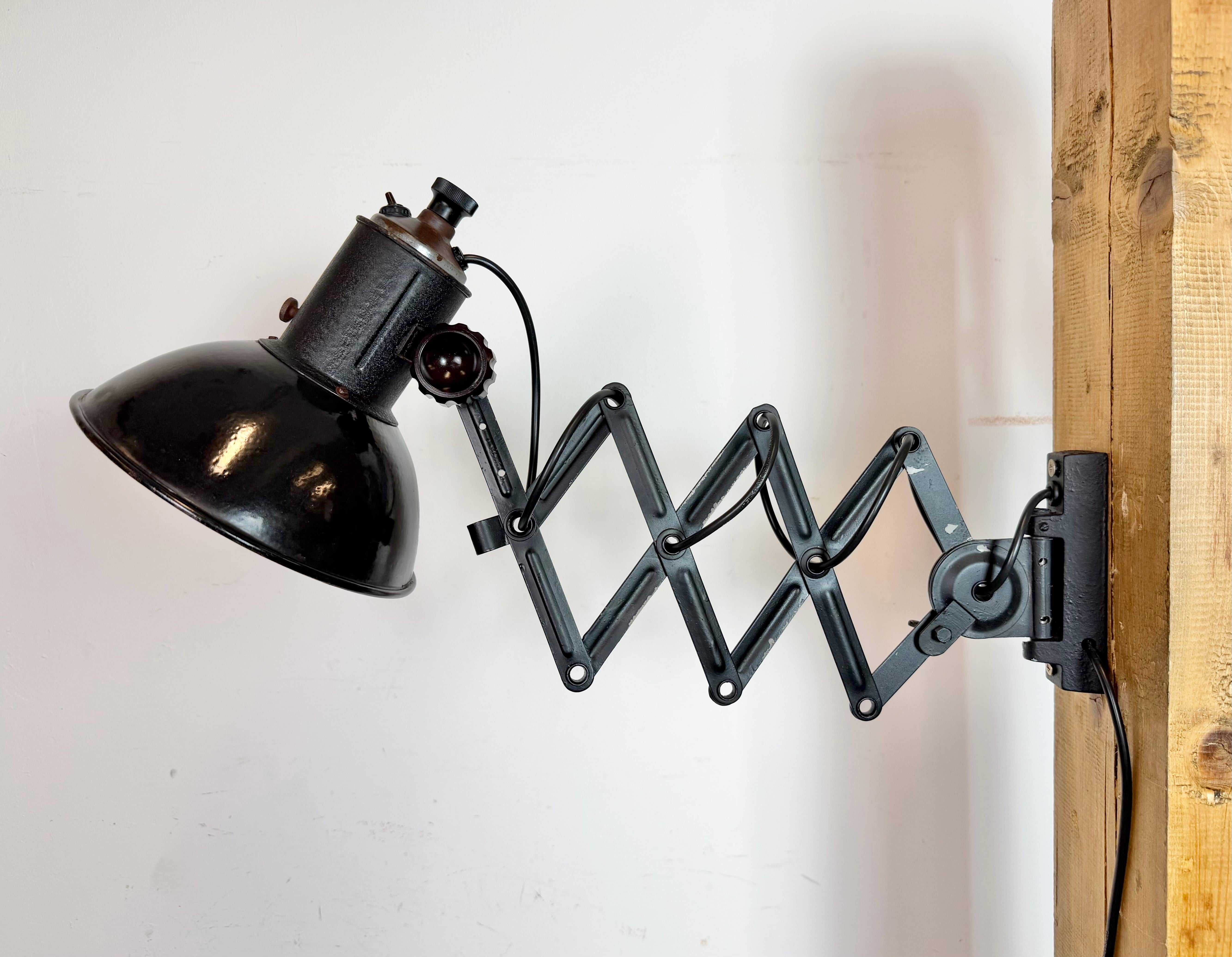 Este aplique industrial vintage de tijera regulable se fabricó en la antigua Checoslovaquia durante la década de 1950. La lámpara tiene una pantalla metálica esmaltada en negro con el interior esmaltado en blanco. Un brazo de tijera de hierro negro