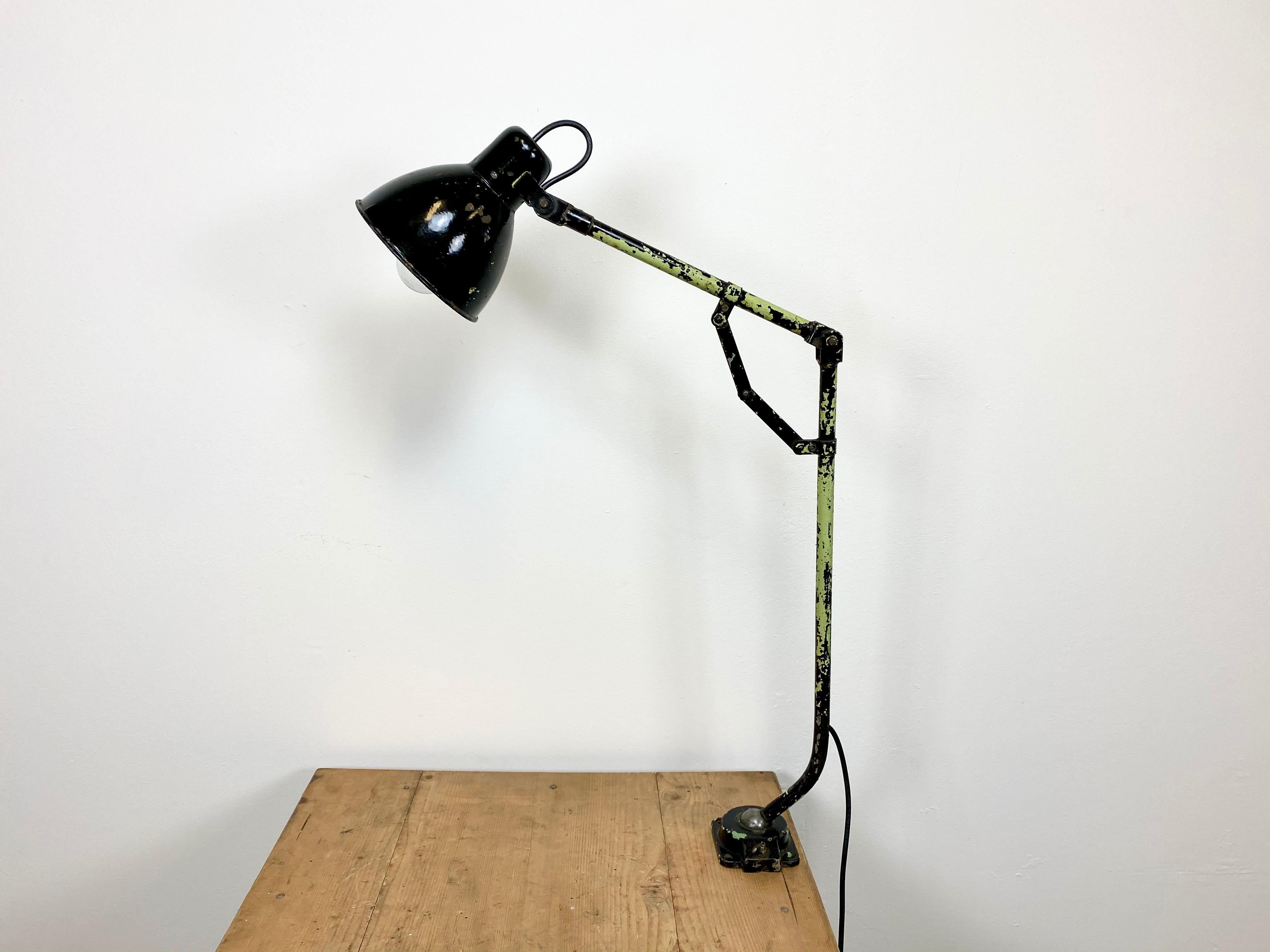 Industrial workshop desk lamp from 1950s. It features a black enamel shade with white interior, an iron base and arm with three adjustable joints. The porcelain socket requires E27 lightbulbs. The diameter of the shade is 16 cm.
