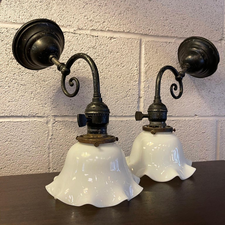 Pair of industrial, wall sconce lamps feature scrolled, blackened nickel stems with scalloped milk glass shades. The lamps are wired to accept a 100 watt bulb each with backplates that measure 4.5 inches diameter.