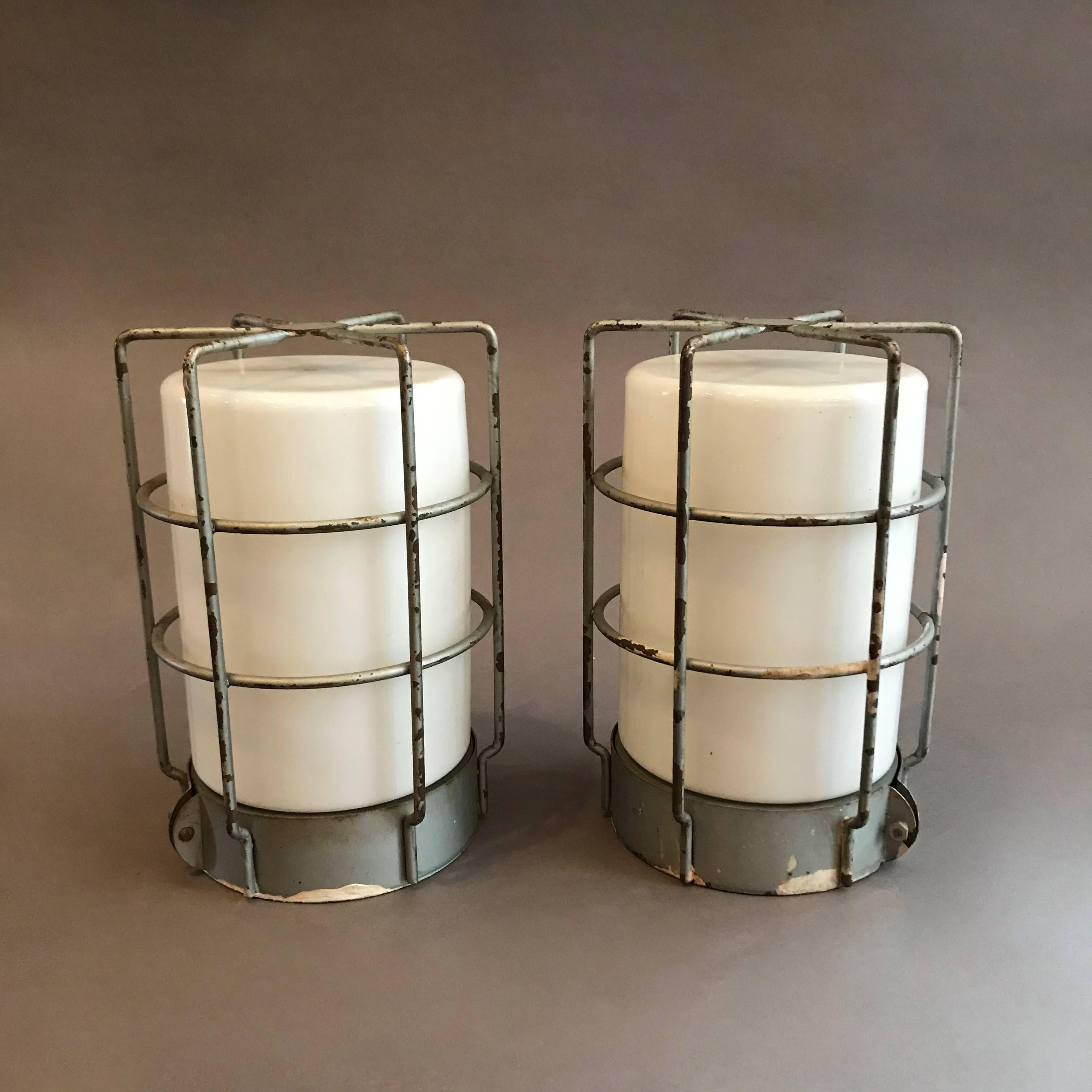 Pair of industrial, flush mount, ceiling light fixtures or wall sconces feature milk glass shades enclosed in steel cages. The fixtures are wired to accept 150 watt bulbs each.