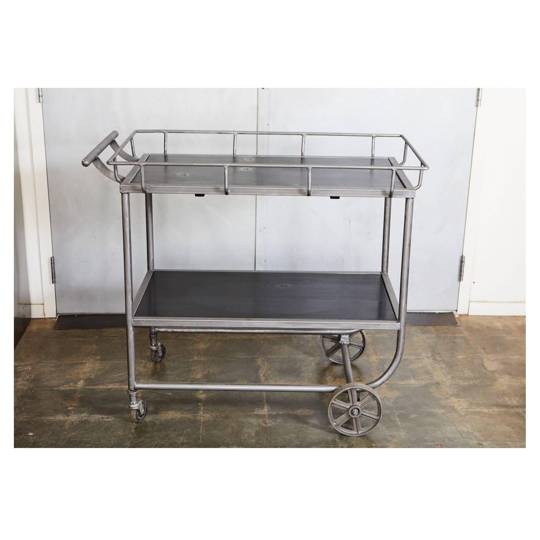 This industrial bar cart has a inset ebony stained wood top and lower shelf. The cart has a push handle, a distinctive gallery made of metal bars and Industrial oversized wheels at the front end making it ideal for entertaining, storage and display