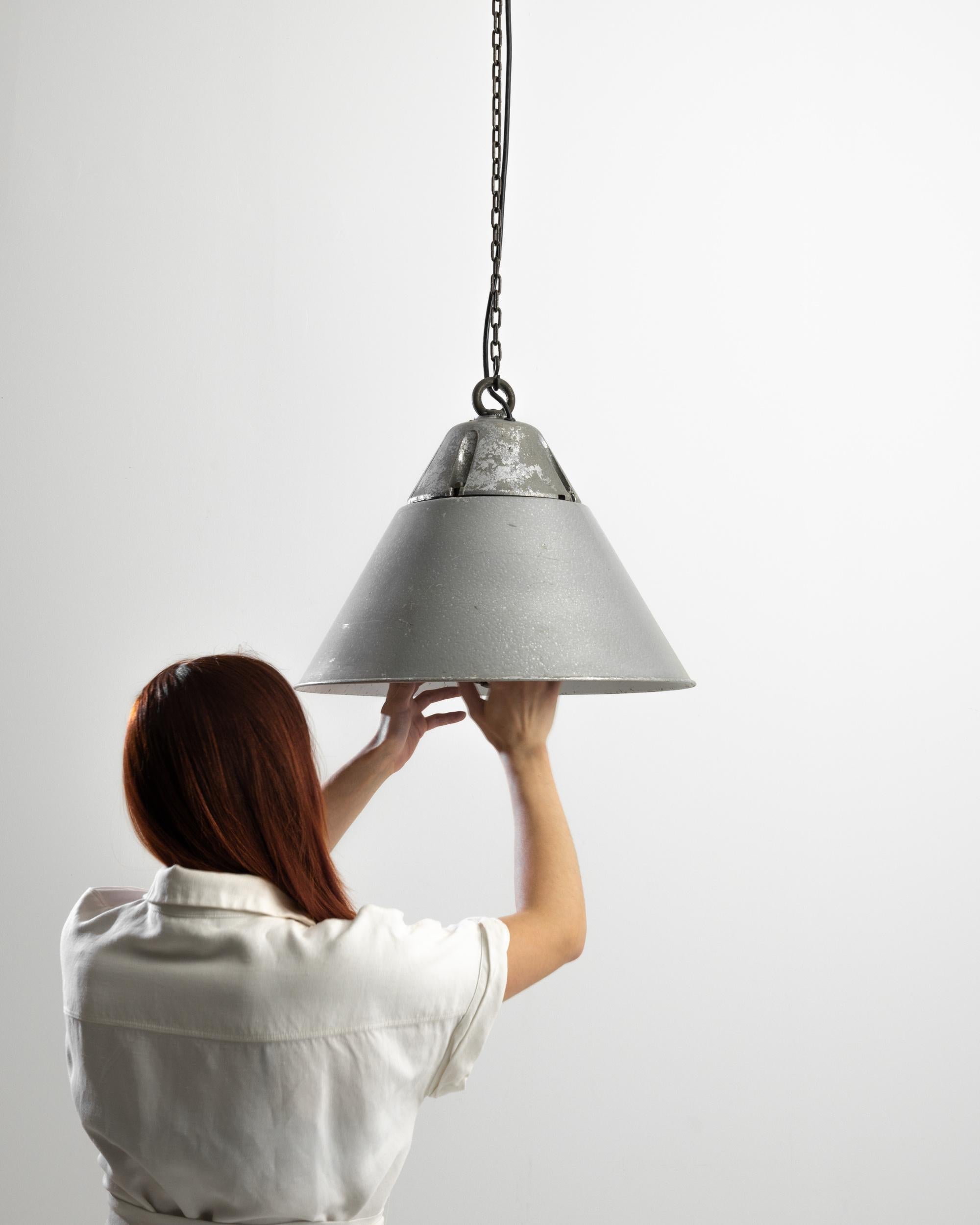Sourced in Central Europe, this 20th Century piece instills a distressed industrial elegance. Suspended on a chain and arrow, the stripped down metal shade exhibits a white interior and a flared profile, creating a diffuse light for an intimate