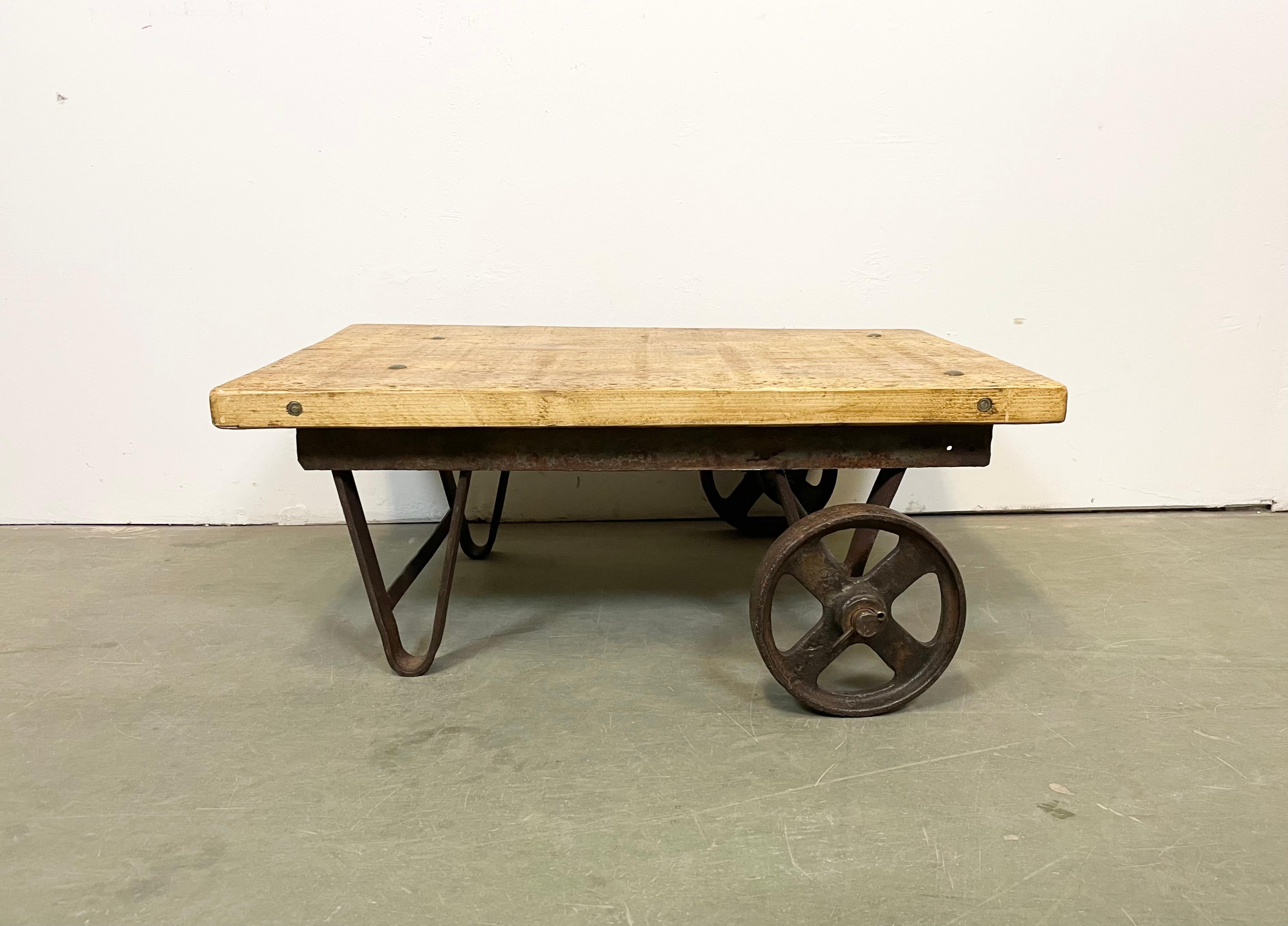 Former pallet truck from a factory now serves as a coffee table. It features a cast iron construction with two original wheels and a solid wooden plate. The weight of the table is 32 kg.