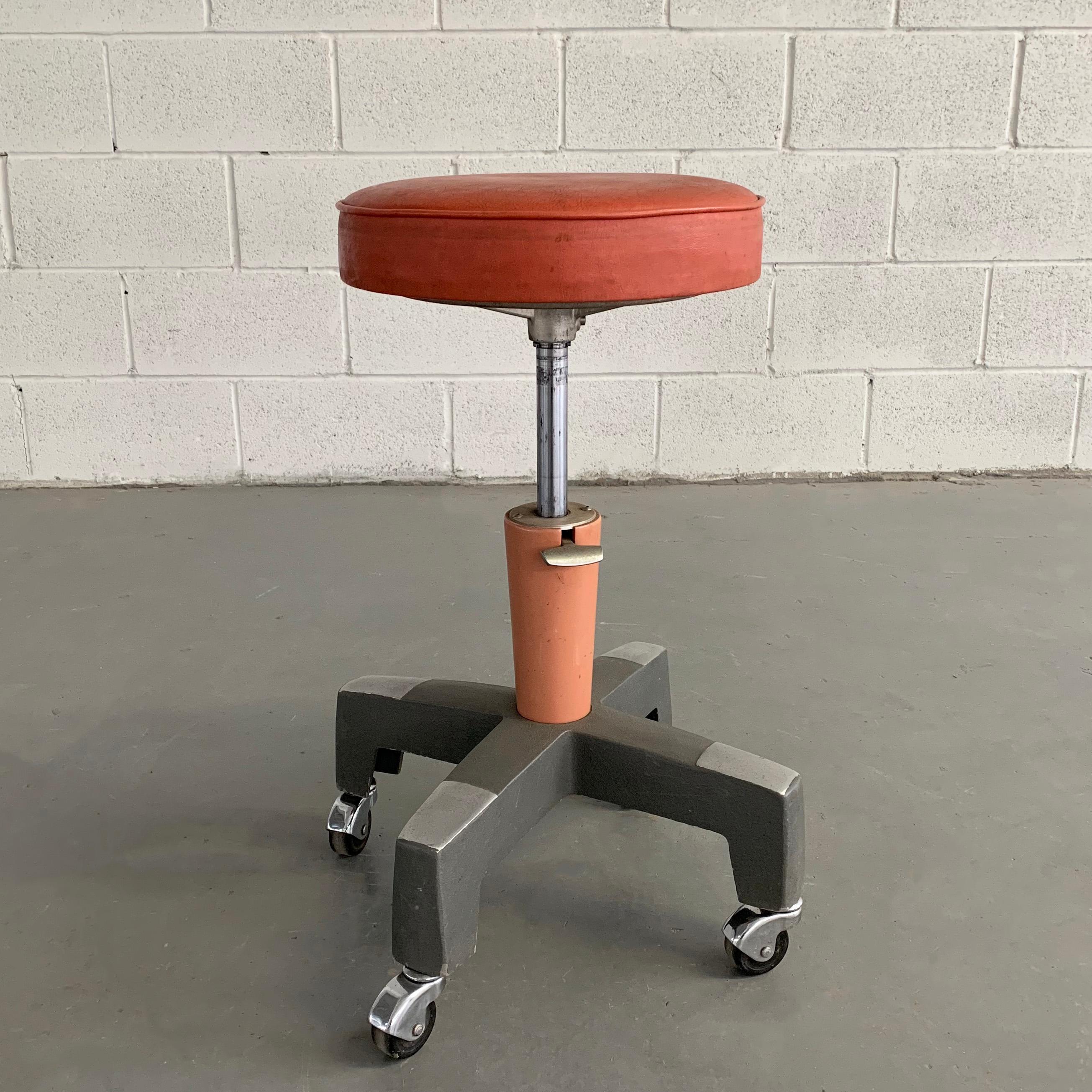 Industrial, midcentury, medical, optometrist stool by American Optical Company with orange vinyl upholstered, 12 inch diameter seat is height adjustable from 17 - 25 inches with enameled base adjustment.