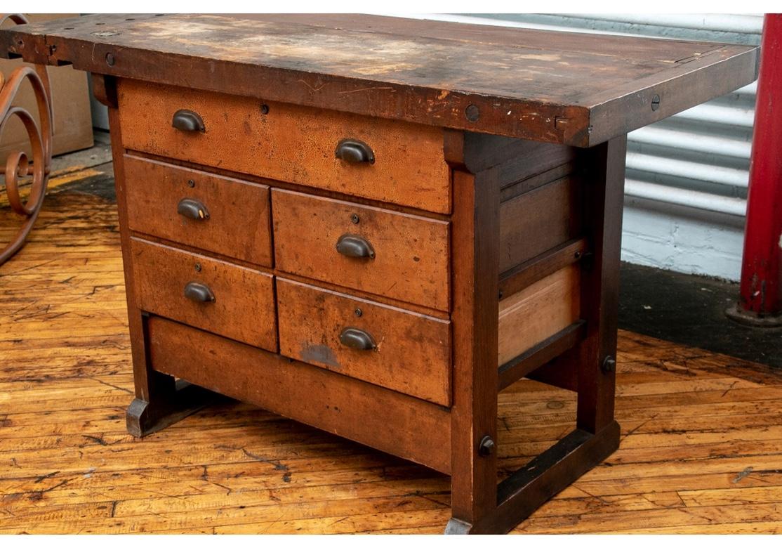 A well used and worn Industrial Era work table made in several parts. With a plank constructed overhanging top with banded ends, the left end with holes. A metal lift up element on top. On a cabinet with a long drawer over two banks of two short