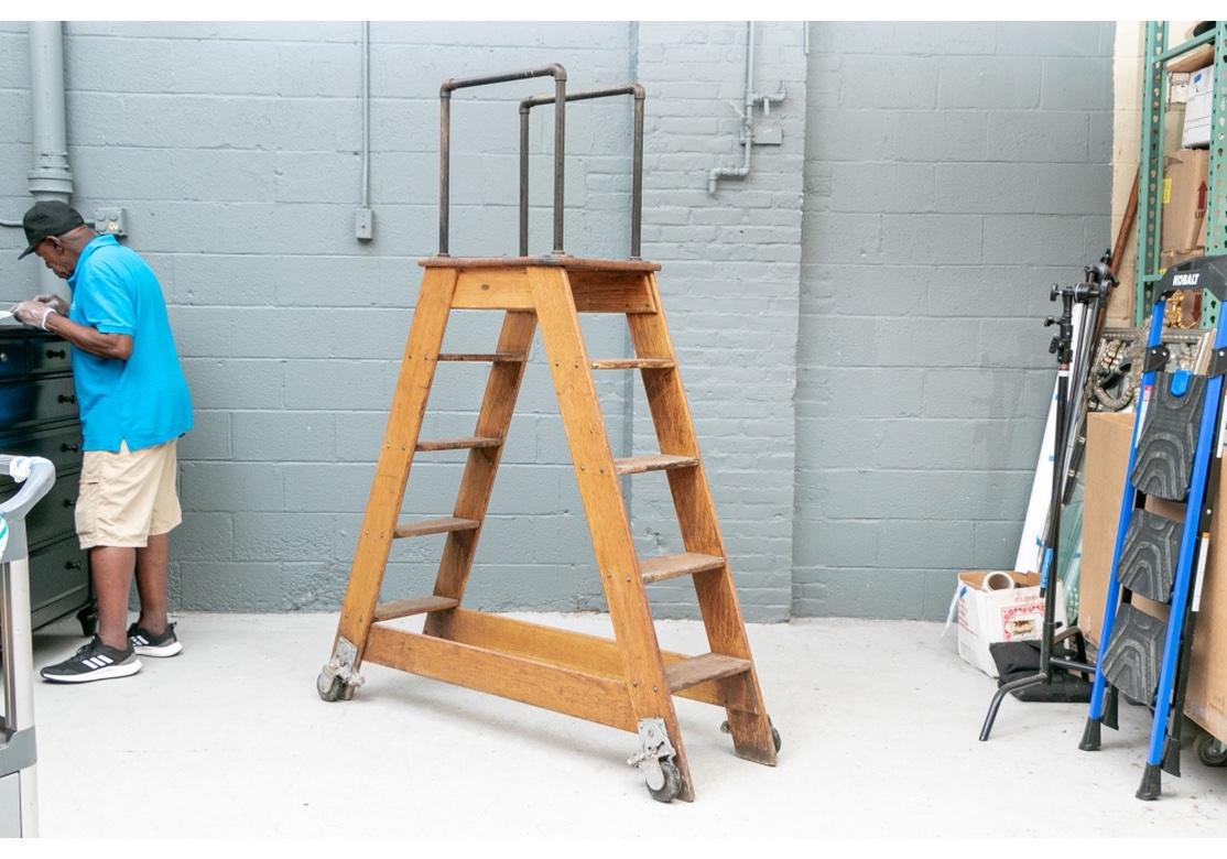 A two- sided four rung ladder with a top mounted with tall open square shaped handles. The front and back connected by flat stretchers. Raised on wheels. Oval Metal number plate affixed. 
Measures: H. 74 1/2