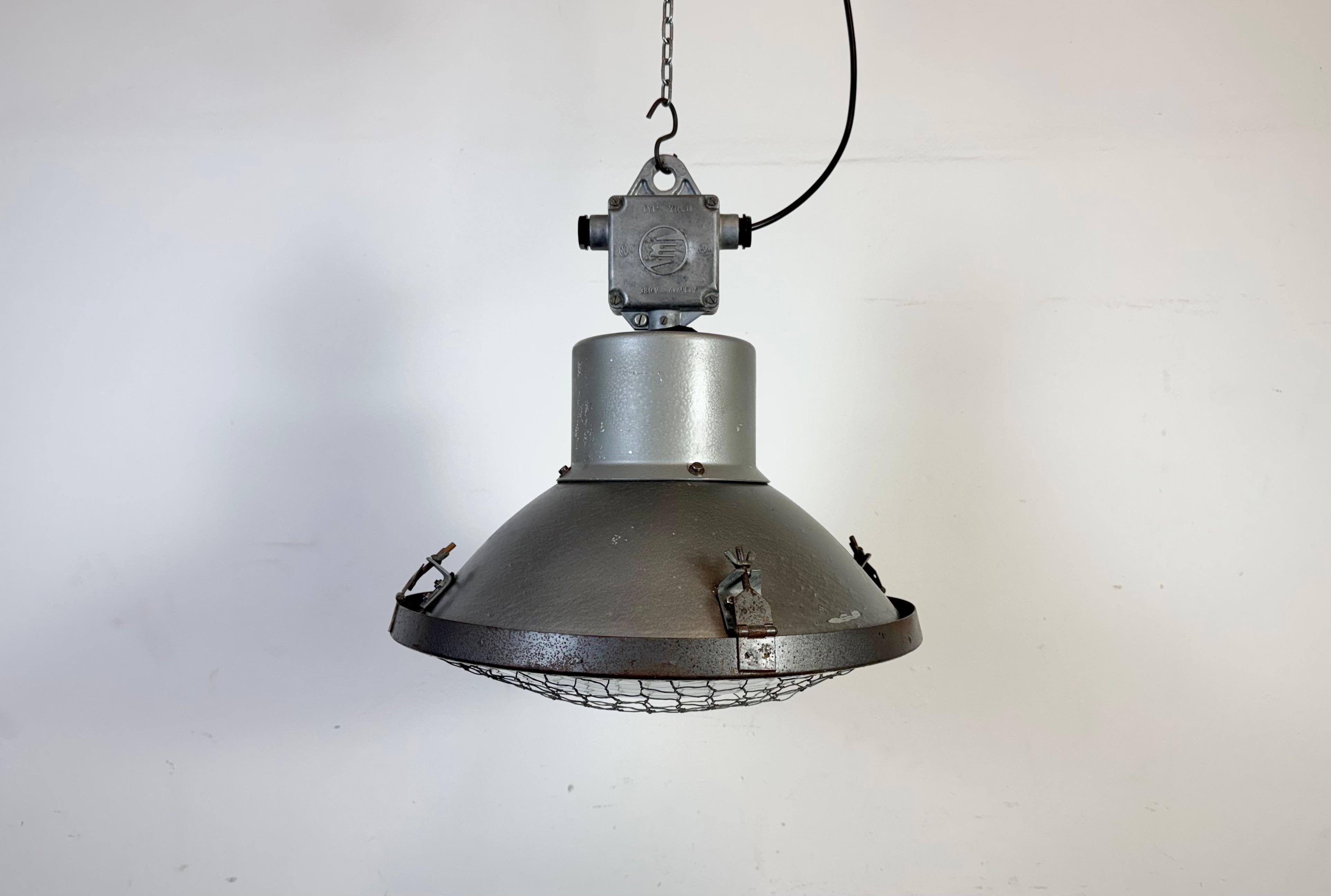 Industrial lamp made by Elektrosvit in former Czechoslovakia during the 1960s It features a grey hammer paint metal body, a cast aluminium top box and a convex clear glass cover with iron grid. The socket  requires a standard E26/E27 light bulbs.