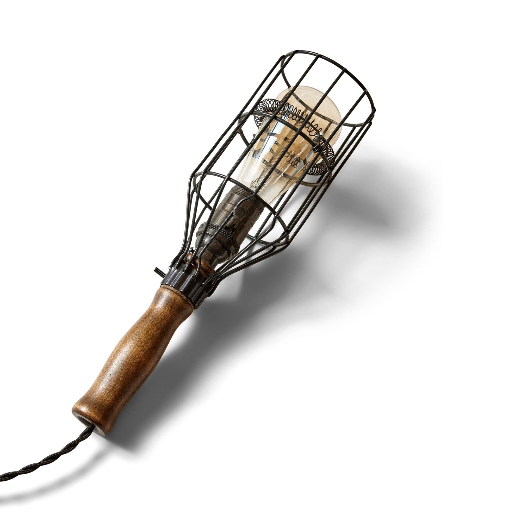 Une lampe utilitaire suspendue en cage de métal noir avec une pince vissée qui la maintient solidement au manche en bois. L'ampoule est soutenue par une bobine métallique.