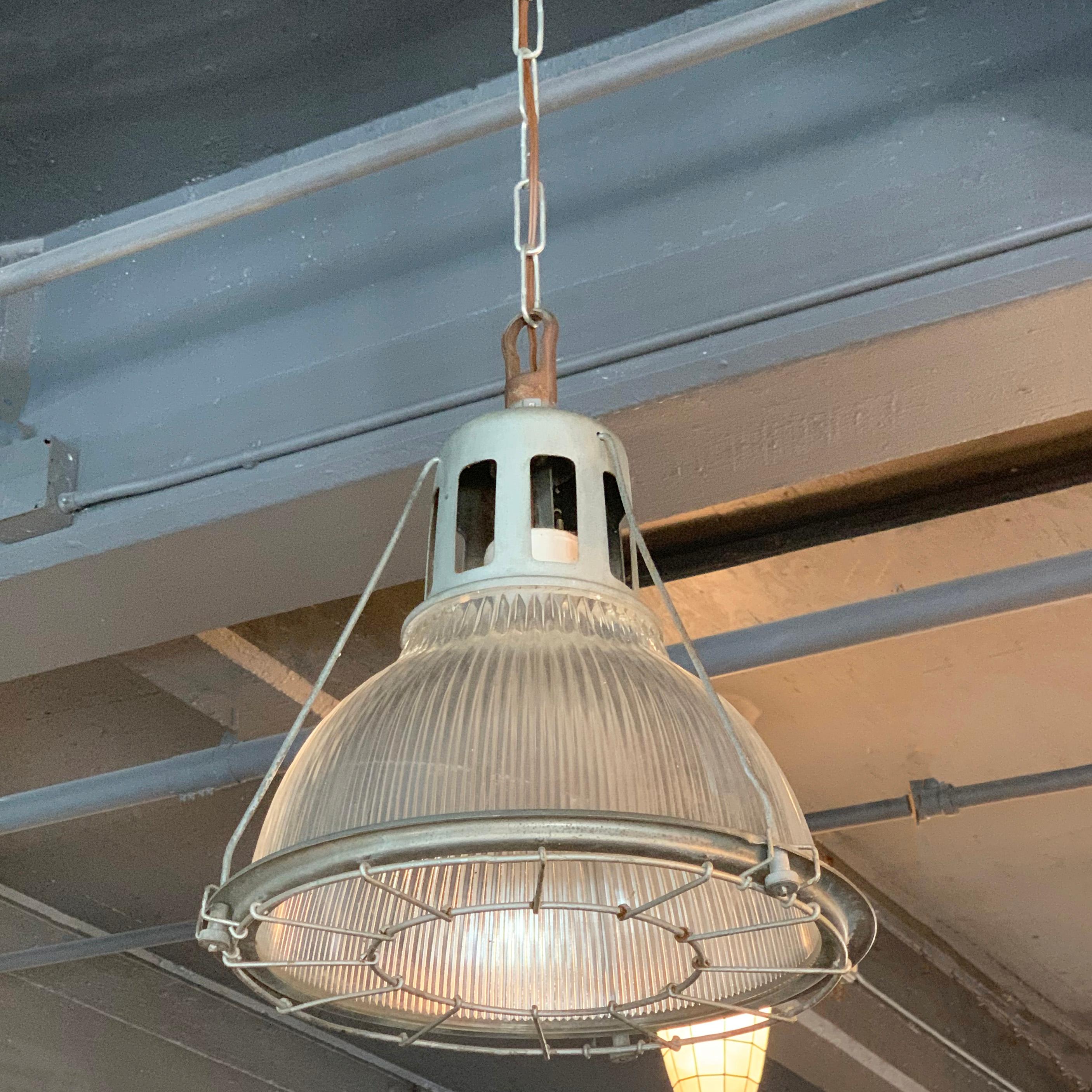 Single, industrial, factory pendant light features a Holophane glass shade within a steel cage frame with porcelain mogul socket. The pendant is newly wired to accept up to a 200 watt mogul bulb or a medium socket bulb with included adaptor and